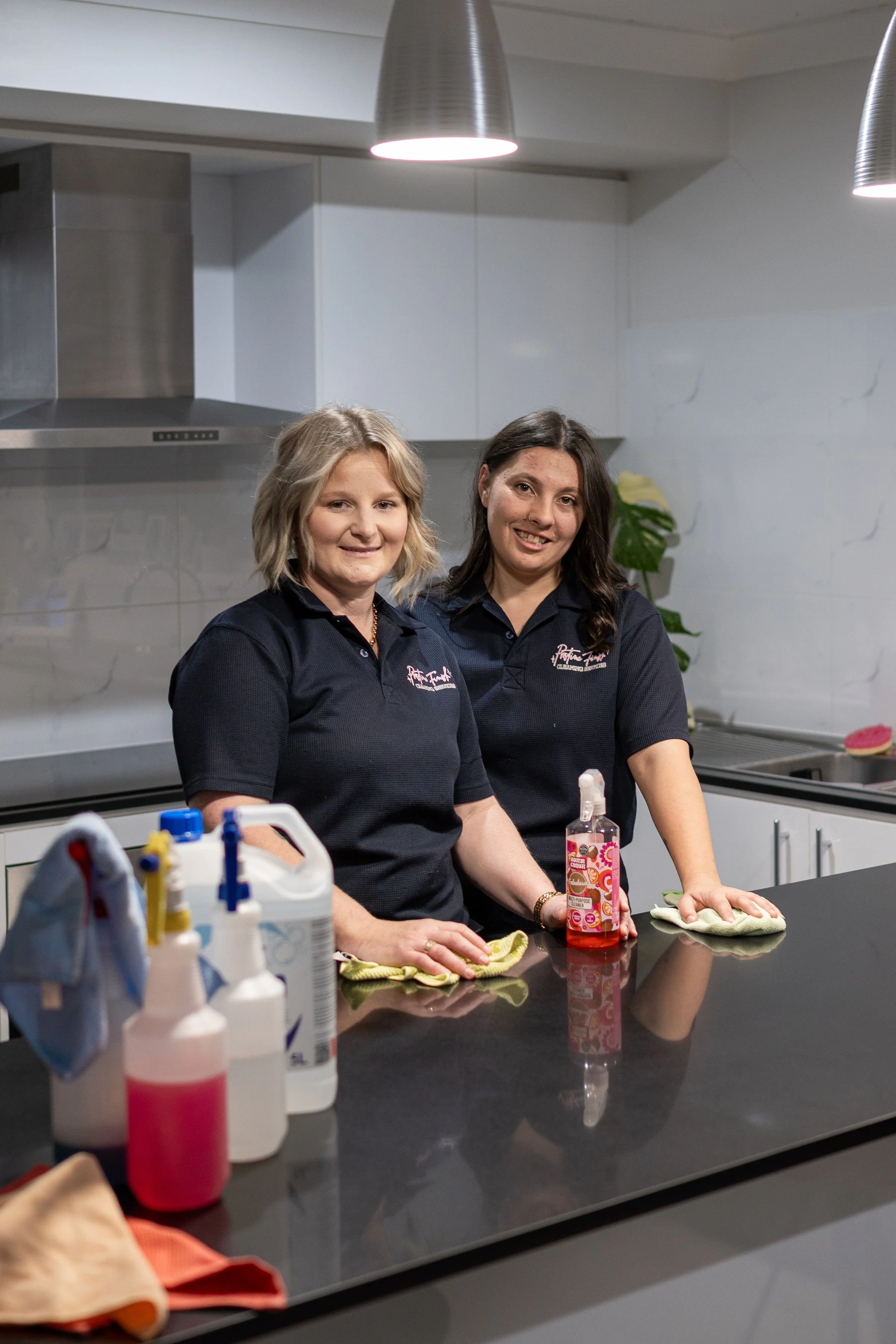 Two women in black shirts cleaning a black countertop in a modern kitchen, with cleaning supplies and a hand sanitizer bottle on the counter.