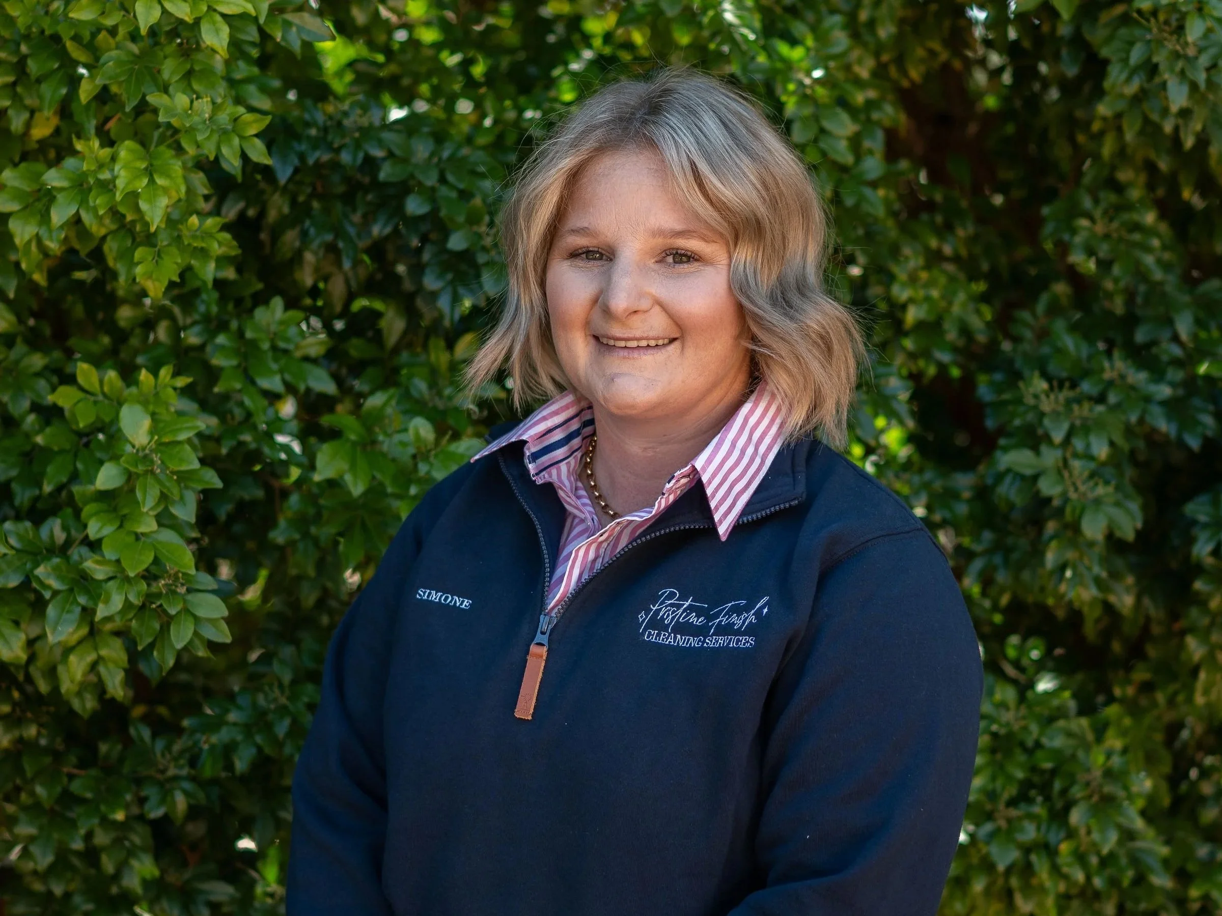 A woman with short blonde hair and a striped shirt underneath a navy jacket with a logo, standing in front of leafy green bushes, smiling at the camera.