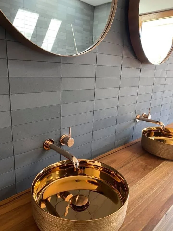 Two gold-colored vessel sinks with matching wall-mounted faucets on a wooden vanity in front of a dark tiled wall, with round mirrors above each sink.