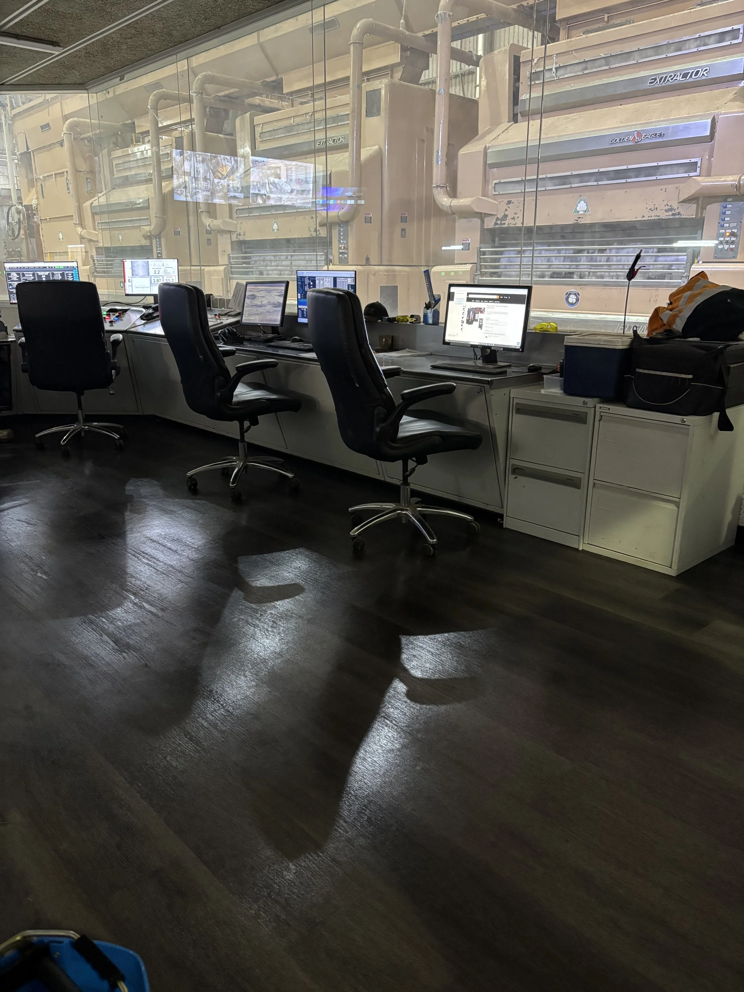 View of an office with three black chairs and multiple computer monitors on a white desk. Behind the desk is a glass window through which large industrial manufacturing machines can be seen.