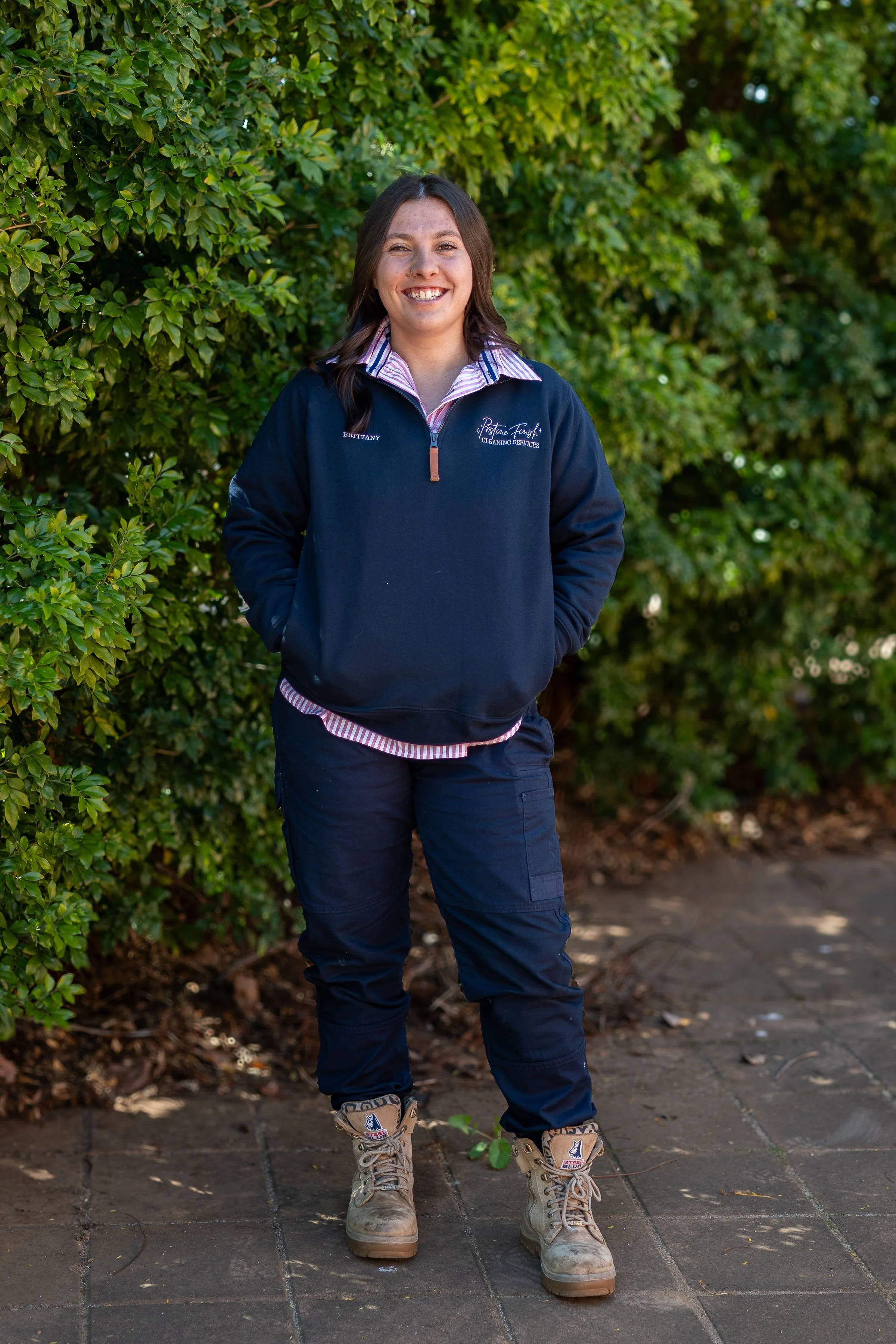 A woman standing outdoors on a brick pathway, smiling, wearing a navy blue uniform with a zipper jacket, cargo pants, and tan work boots, surrounded by green foliage.