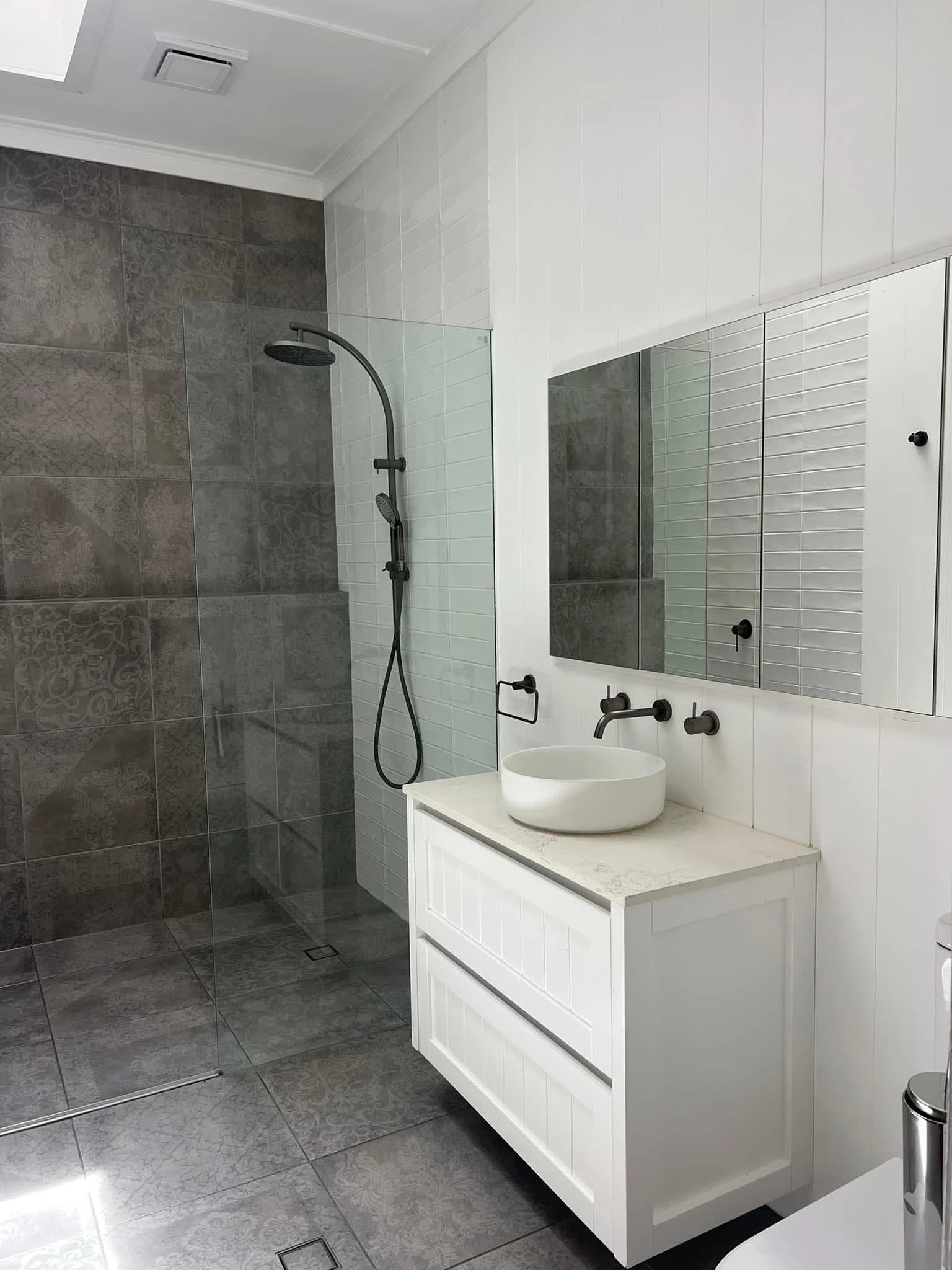 New build move in cleaning Modern bathroom featuring a walk-in shower with dark patterned tiles and a matte black showerhead, a white vanity with a vessel sink, a large mirror, and white tiled walls.
