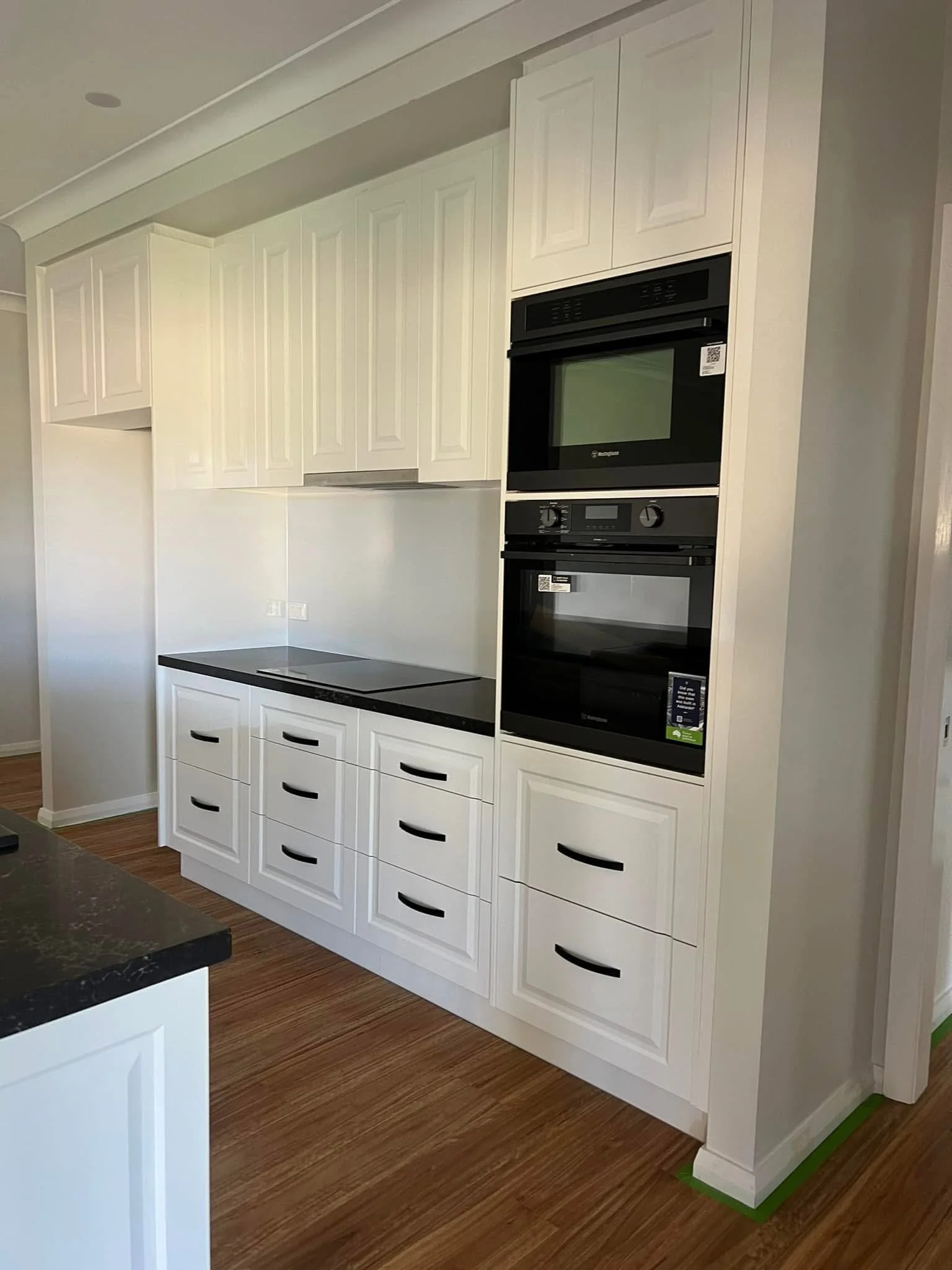 White kitchen cabinets with black handles, a black countertop, and built-in black oven and microwave, with wooden flooring.