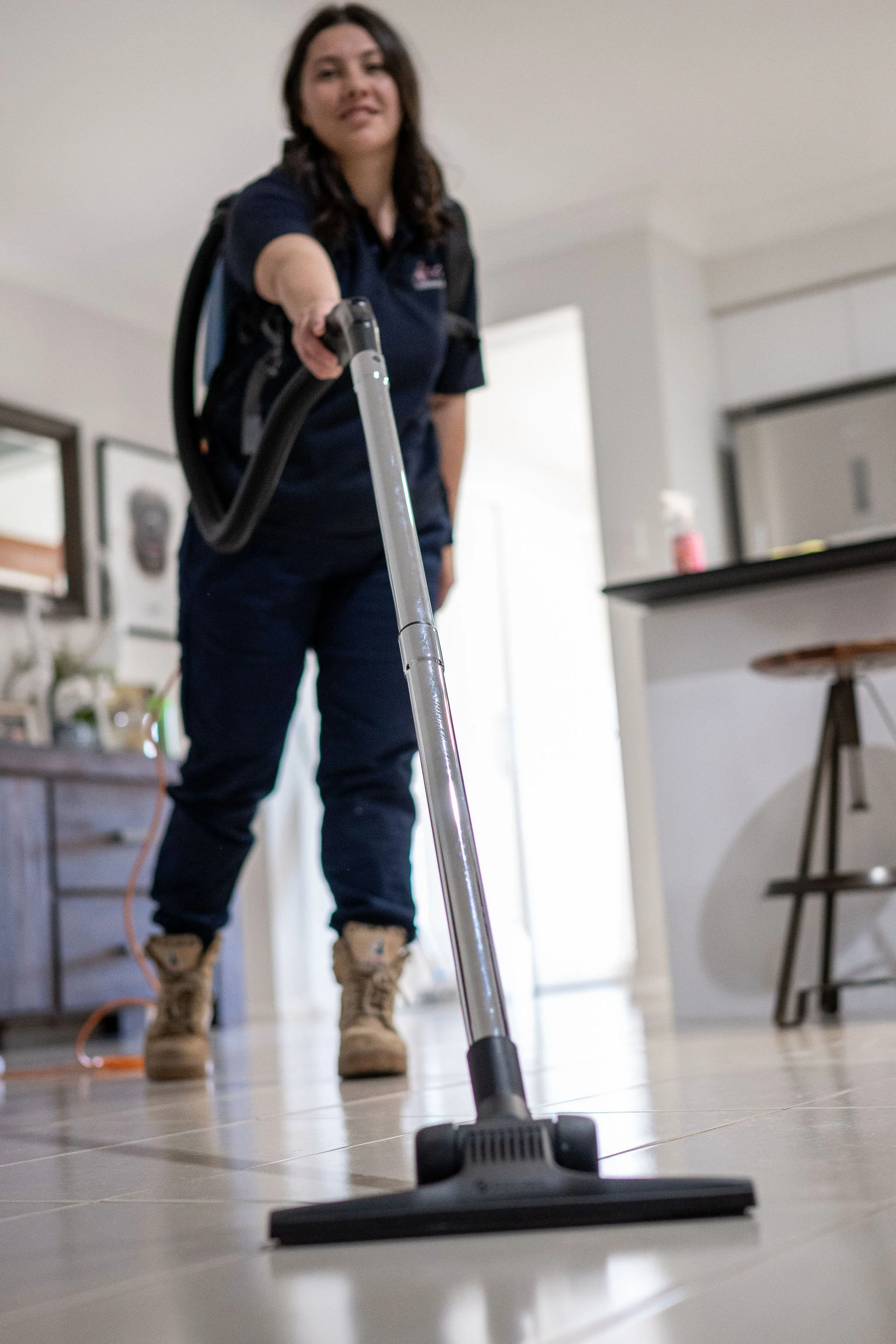 A woman wearing work boots and casual clothing vacuuming a tiled floor in a home interior. Tottenham cleaning company