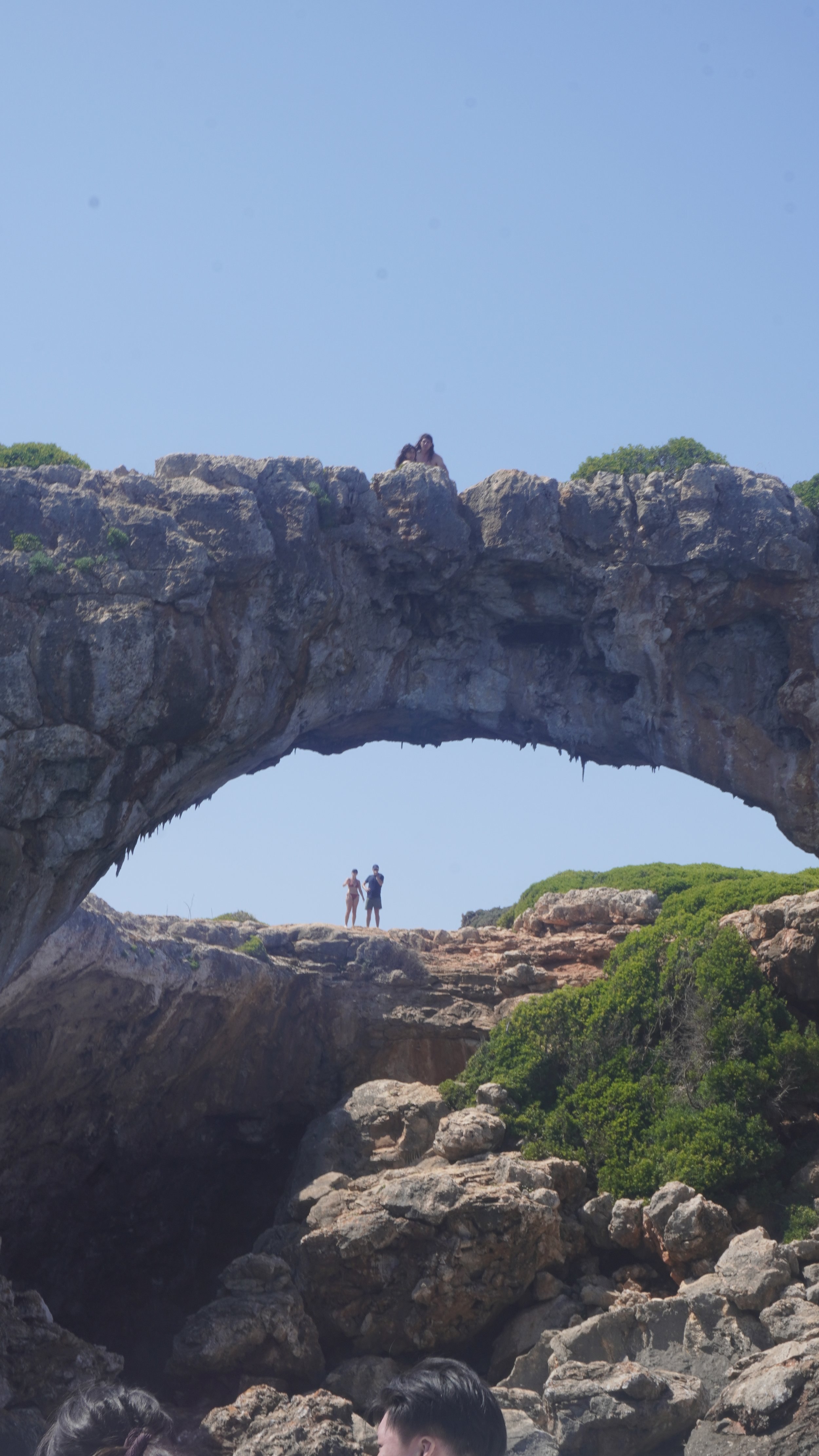 People standing on a large rock formation with a natural stone arch and a person sitting on top of the arch, under a clear blue sky.