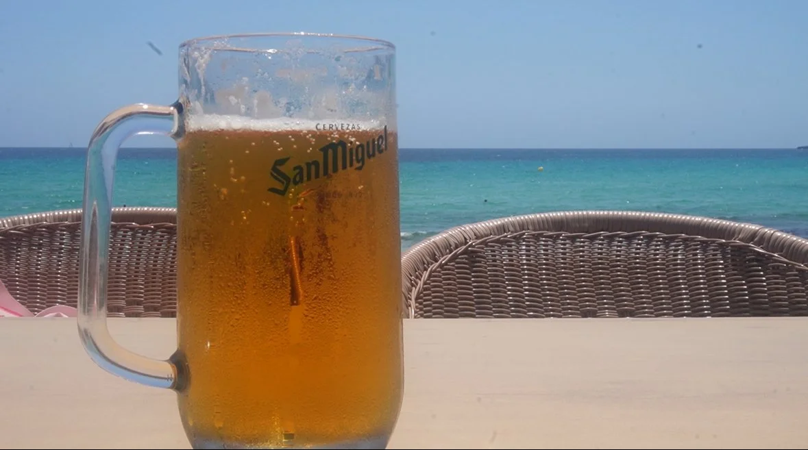 A glass of beer with bubbles, placed on a table near the beach with wicker chairs and ocean in the background.