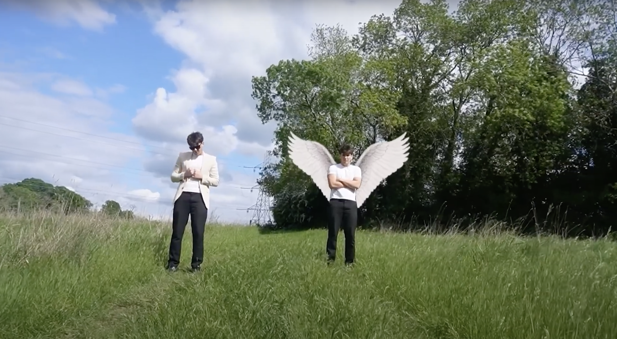 Two young men standing outdoors in a grassy field, one with white angel wings and the other dressed formally, under a partly cloudy sky with trees in the background.
