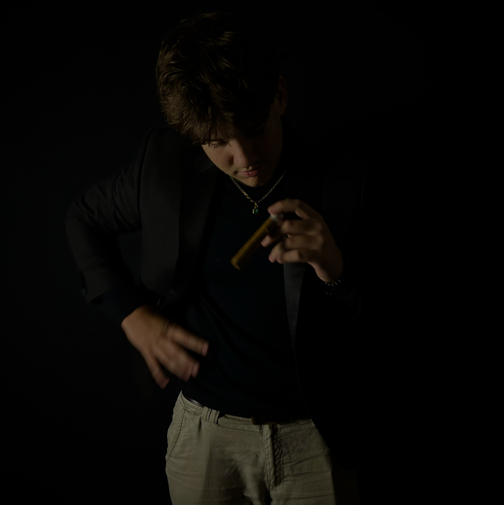A young man with brown hair wearing a black blazer, black shirt, and khaki pants, holding a cigar and looking down against a black background.