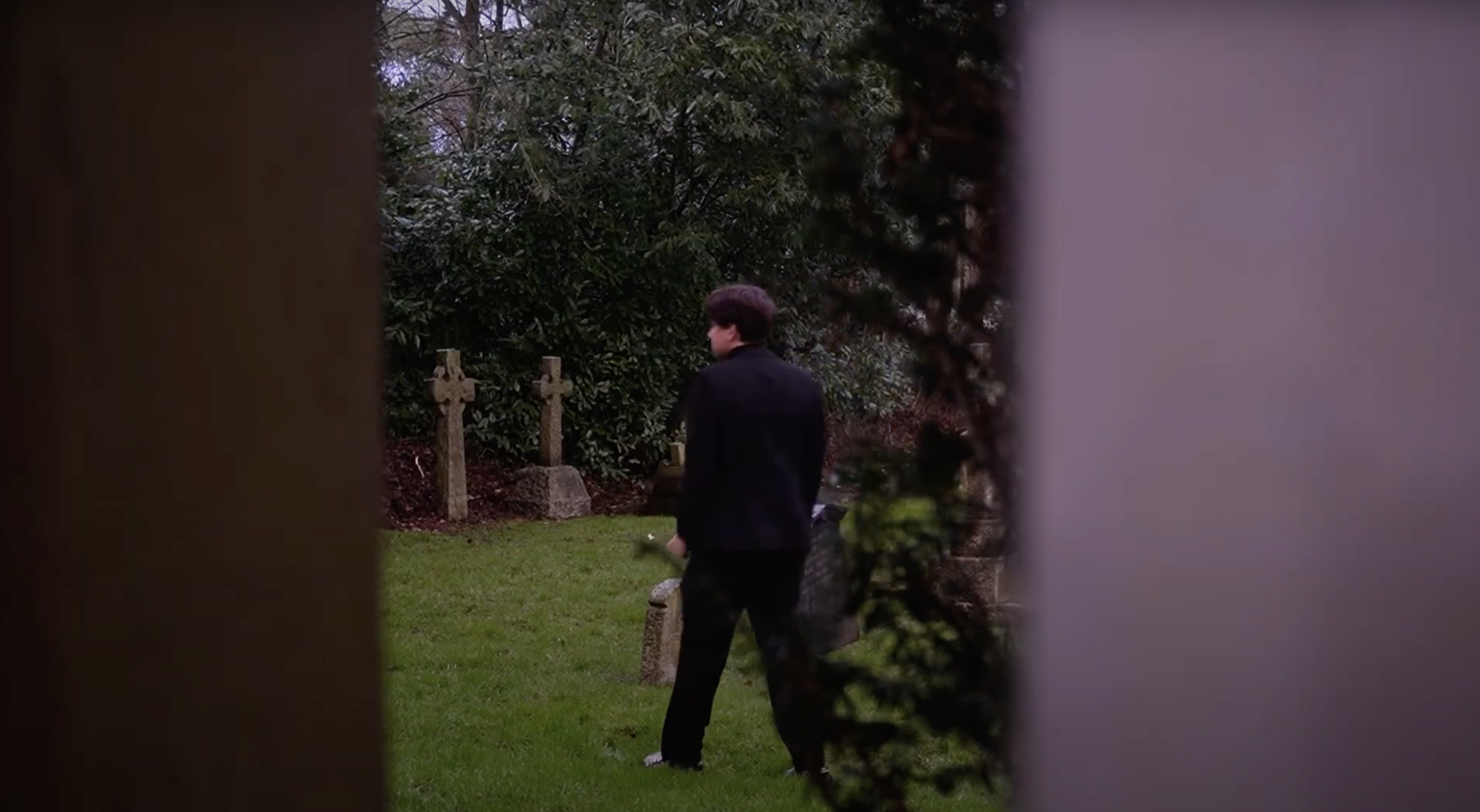 A man dressed in black is walking in a grassy area, seen through a narrow gap between two structures, with trees and crosses in the background.