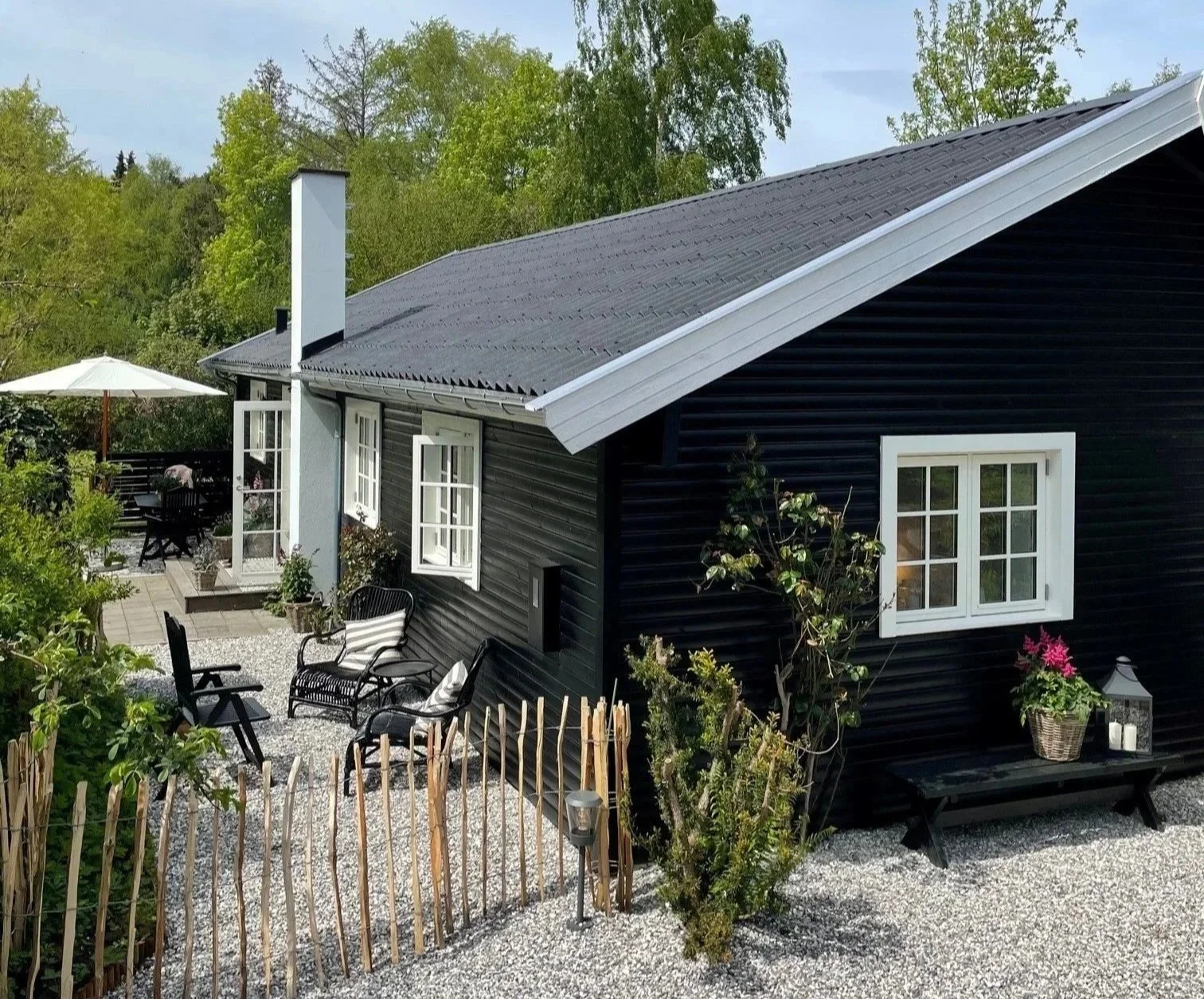 Black wooden house with white-framed windows and a sloped dark gray roof, surrounded by greenery, outdoor seating area with chairs, a table with an umbrella, and a gravel yard.