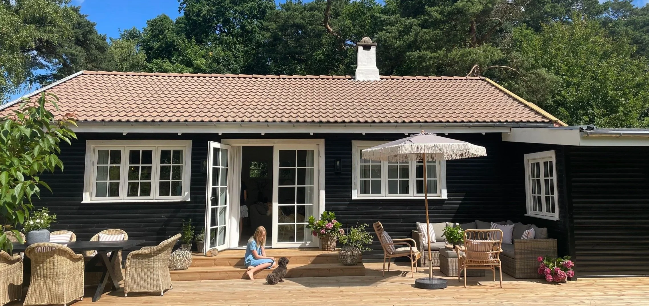 Backyard deck with outdoor seating, large colorful umbrellas, potted plants, a girl and a dog sitting on the steps of a black wooden house with white window frames and a tiled roof, surrounded by trees.