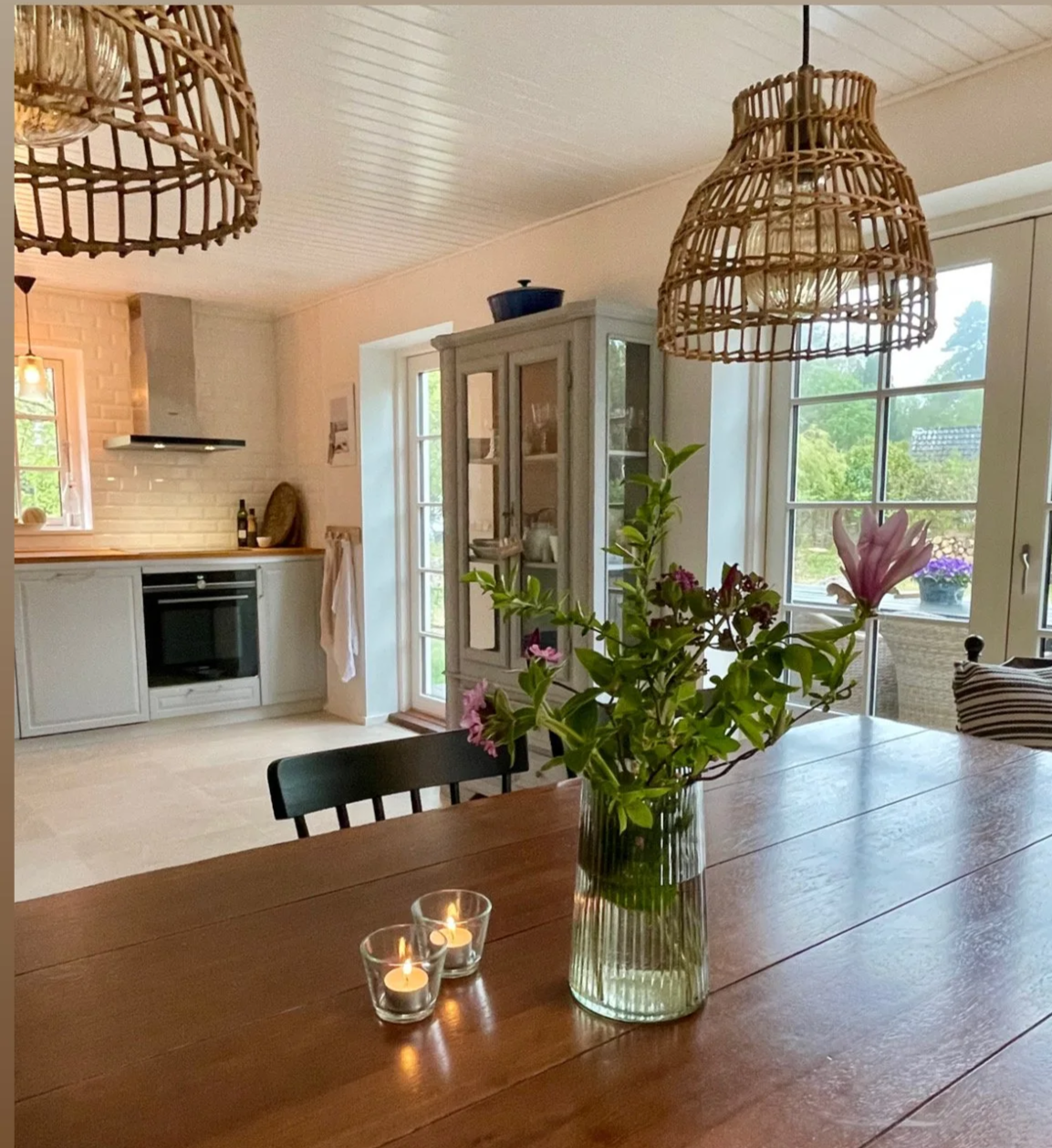A cozy kitchen and dining area with a wooden table, a vase of flowers, and candles, featuring large windows with a view of green trees outside. The kitchen includes white cabinetry, a built-in oven, and hanging wicker pendant lights.