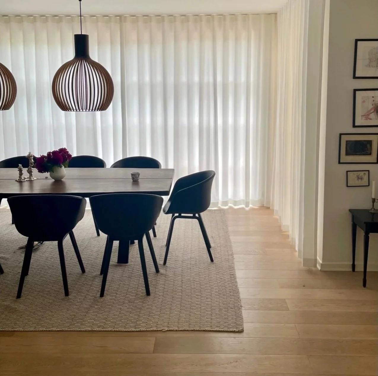 Dining room with a wooden table, six black chairs, a vase of pink flowers, and modern pendant lights. Floor is hardwood with a beige rug. Large sheer curtains cover the windows.