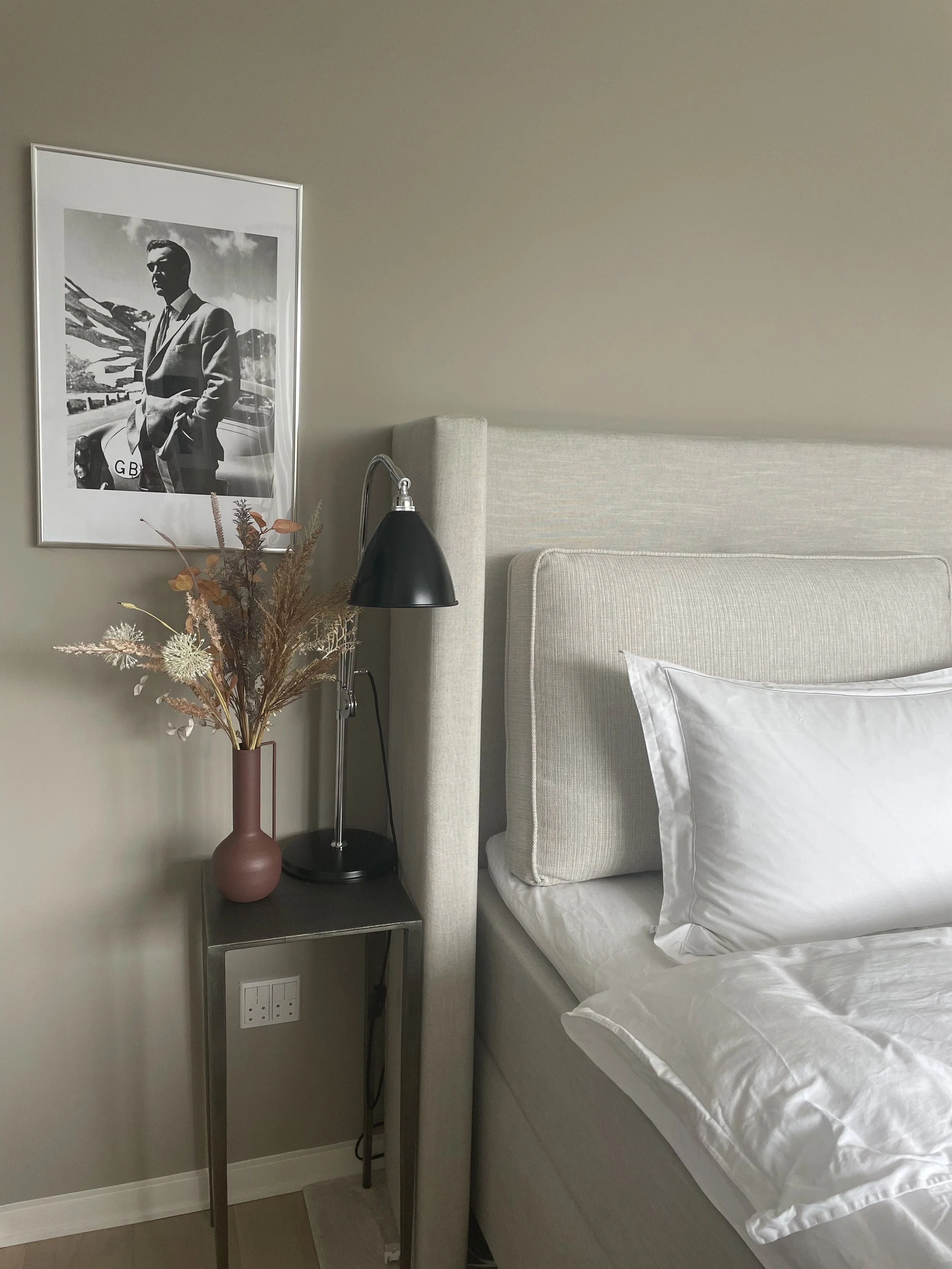 A bedroom corner with a beige upholstered bed, white pillow and bedding, a small side table holding a black desk lamp, a pink vase with dried flowers, and a black-and-white framed photograph of a man in a suit on the wall.