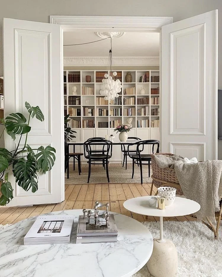 Living room view looking into a dining area with a bookshelf, black dining chairs, and a white table with a flower arrangement. The foreground has a marble coffee table, a side table with cups and decor, and a wicker chair with a blanket.