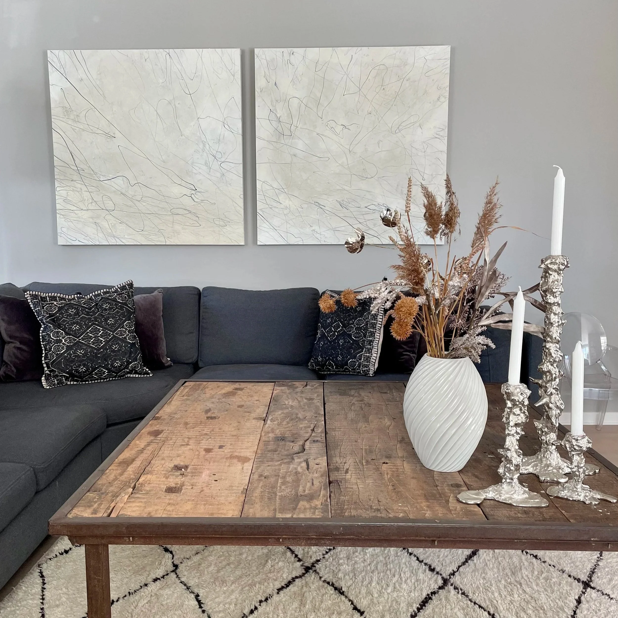 A modern living room with a dark grey sectional sofa, decorative black and silver pillows, a large white vase with dried flowers, and silver candlesticks with white candles on a wooden coffee table, with abstract art on the wall behind.
