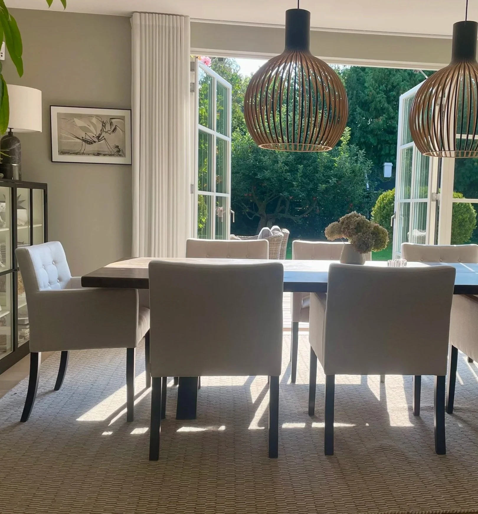 A dining room with a large table, six white upholstered chairs, two pendant lights hanging above, and open glass doors leading to a garden with trees and lawn.