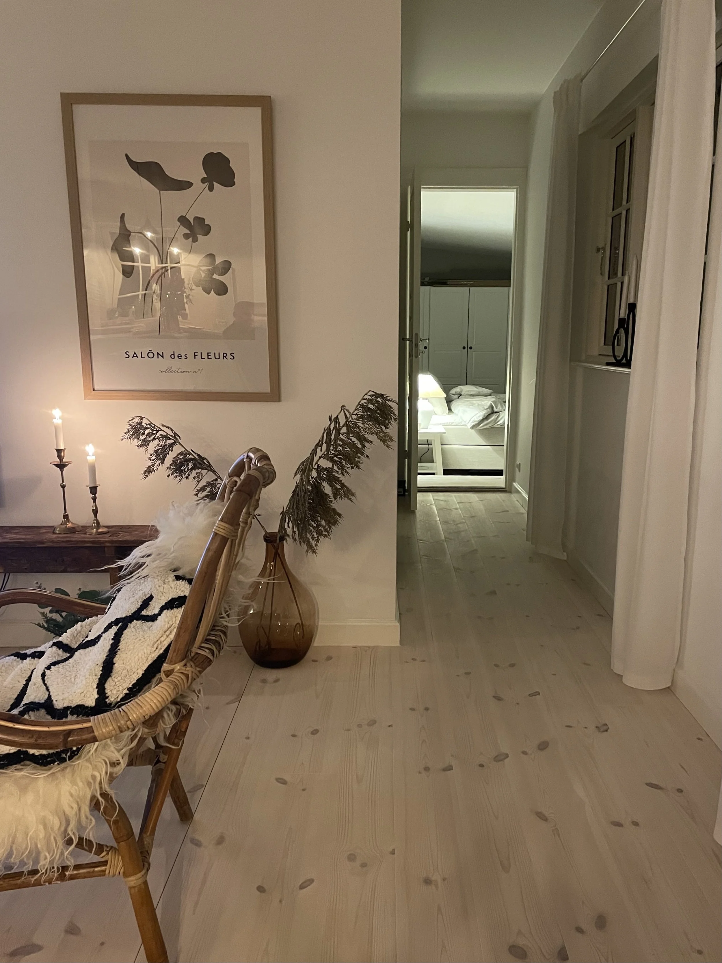 A cozy interior room with a rattan chair covered with a black and white blanket, a brown glass vase holding dried branches, a picture of flowers on the wall, and a hallway leading to a bedroom with a bed and nightstand.