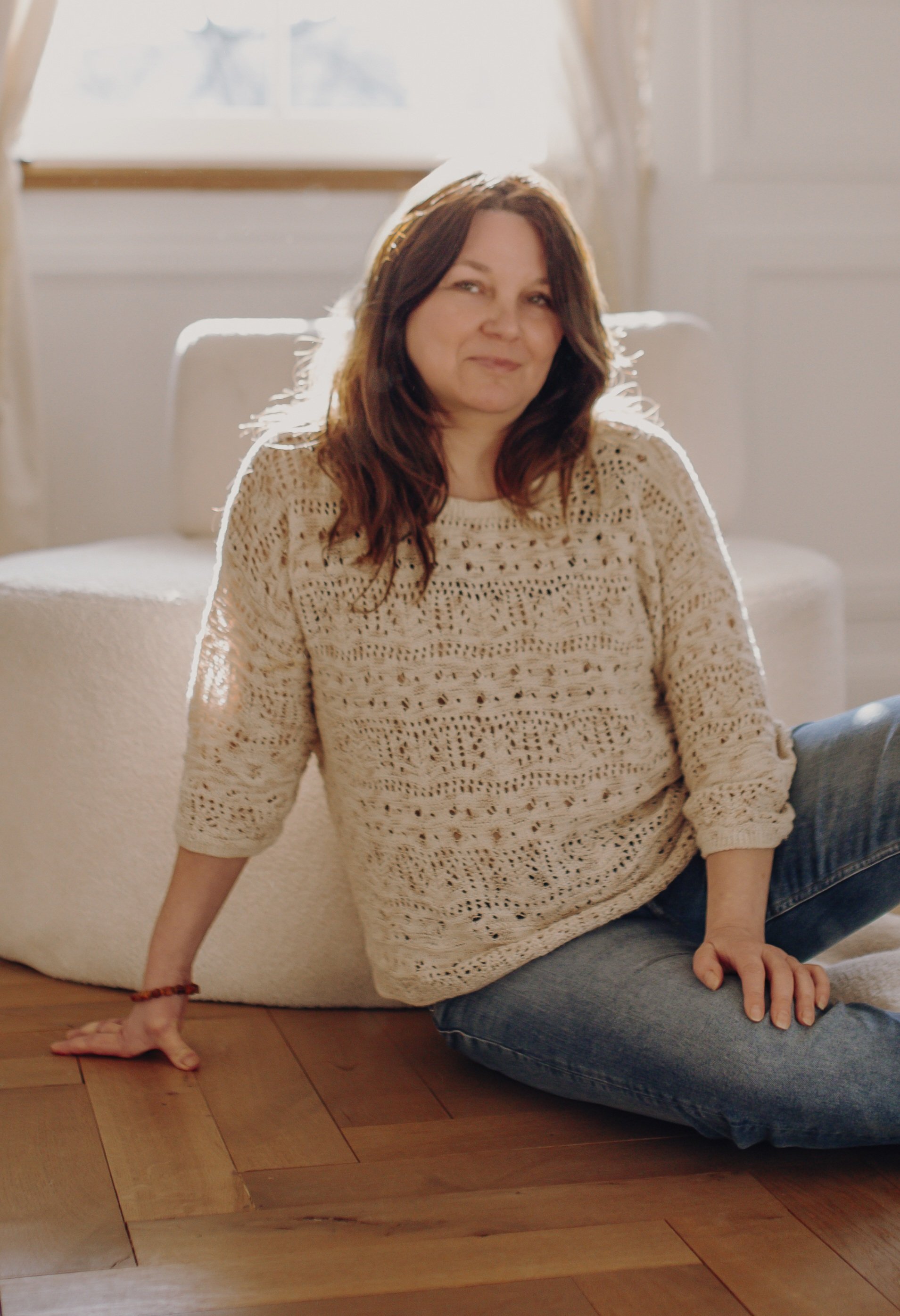 Frau sitzt auf dem Holzboden neben einem weißen, bauschigen Sitzsack, in einem hellen Raum mit Fenster im Hintergrund, Sonnenlicht strahlt auf sie