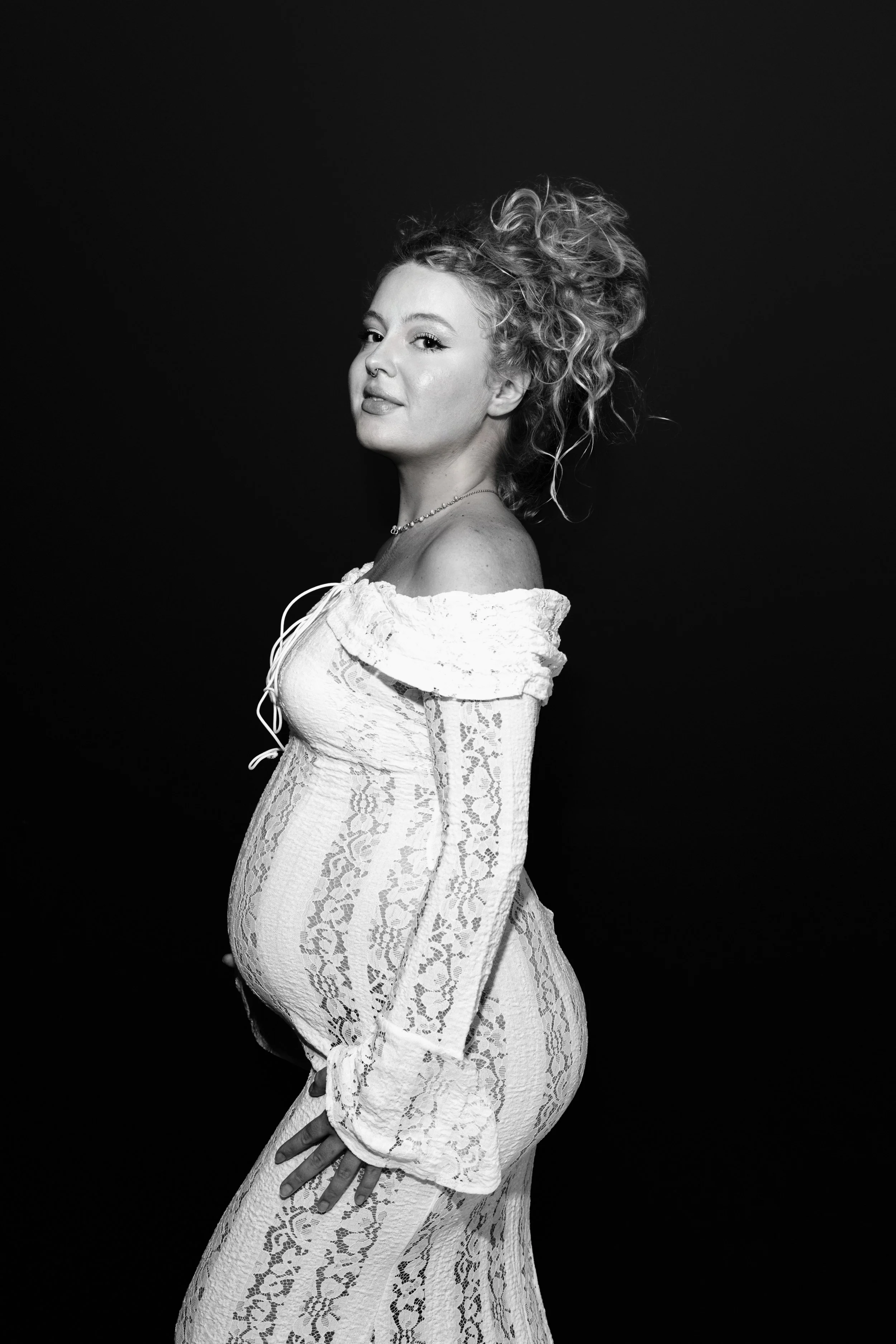 Femme enceinte portant une robe en dentelle blanche, debout de profil, avec un fond noir.