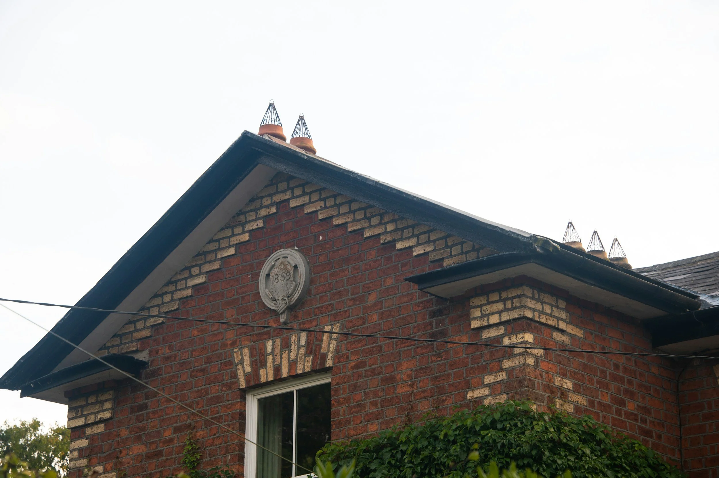 Close-up of the gable end of a red brick house with two chimneys with wire covers, a circular decorative plaque marked '1859', and a window with a white frame and curtain, partly covered by green foliage.