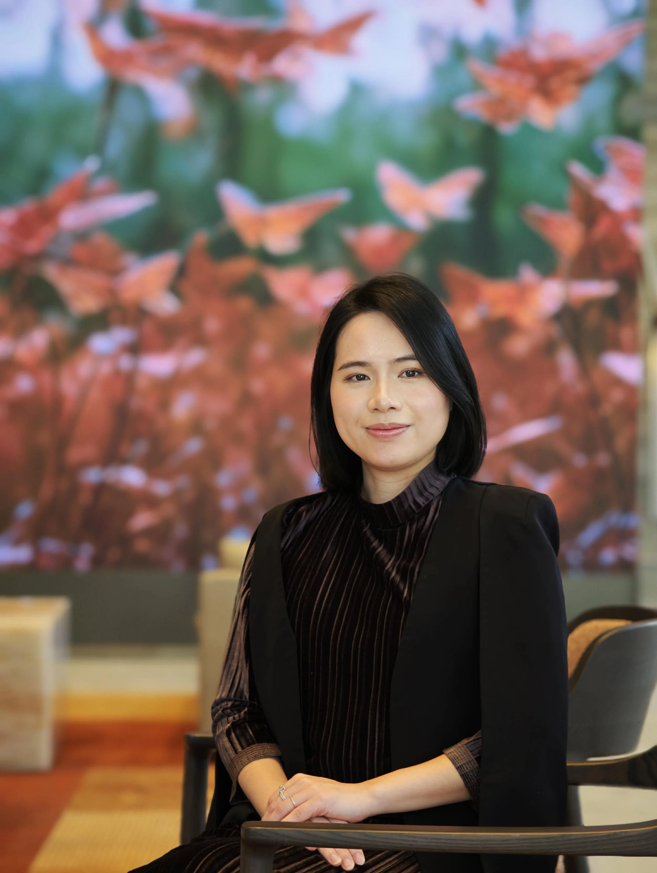 Portrait of an Asian woman with black hair, wearing a black blazer and a striped brown shirt, sitting at a table in a restaurant or cafe with a blurred colorful background.