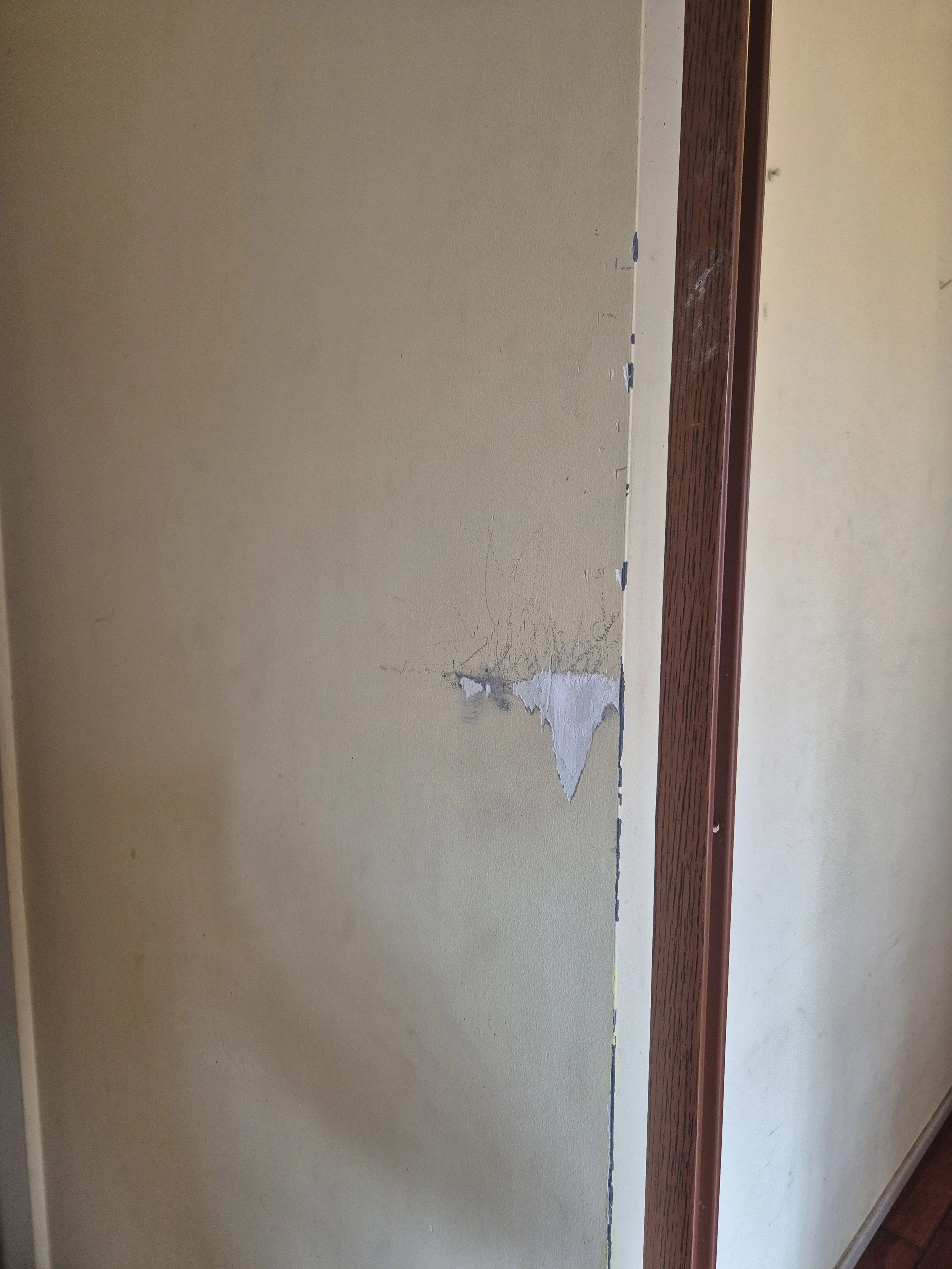 A beige wall with peeling paint and cracks, next to a wooden rail or ledge.