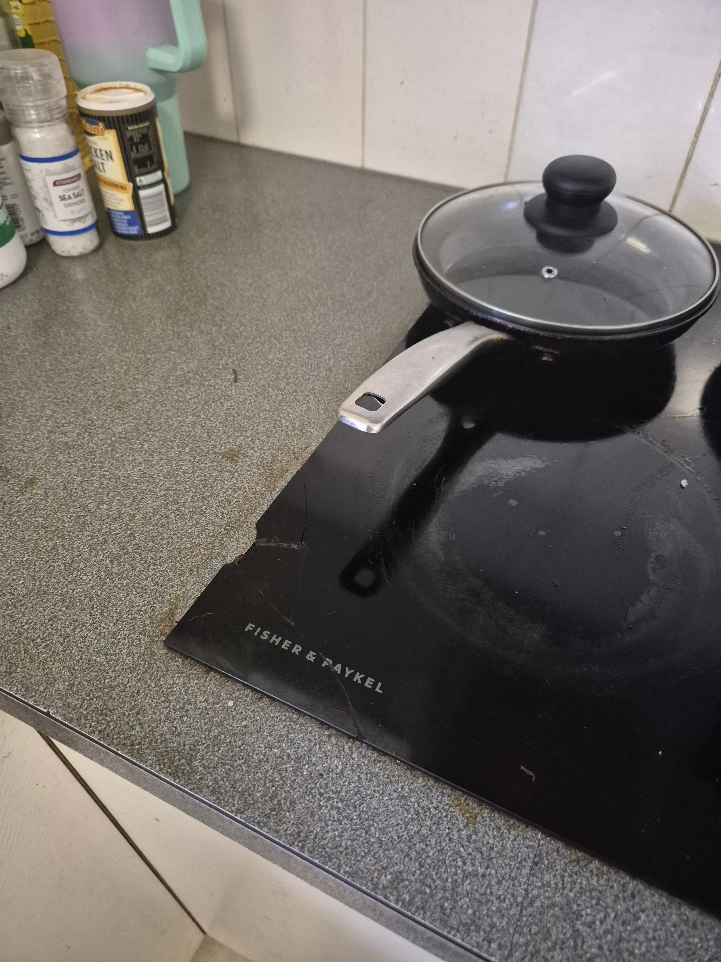 A small frying pan with a glass lid on a black stovetop. The stovetop is on a gray kitchen counter with various spice bottles nearby. The counter has a speckled pattern. The pan is positioned in front of a black mat that says "FISHER & PAYKEL."
