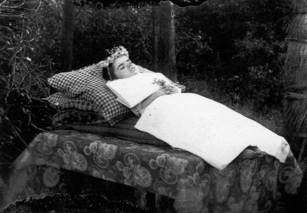 Een vrouw ligt op een bed buiten, met een bloemenkrans op haar hoofd en een witte dekens, omgeven door planten en bomen.