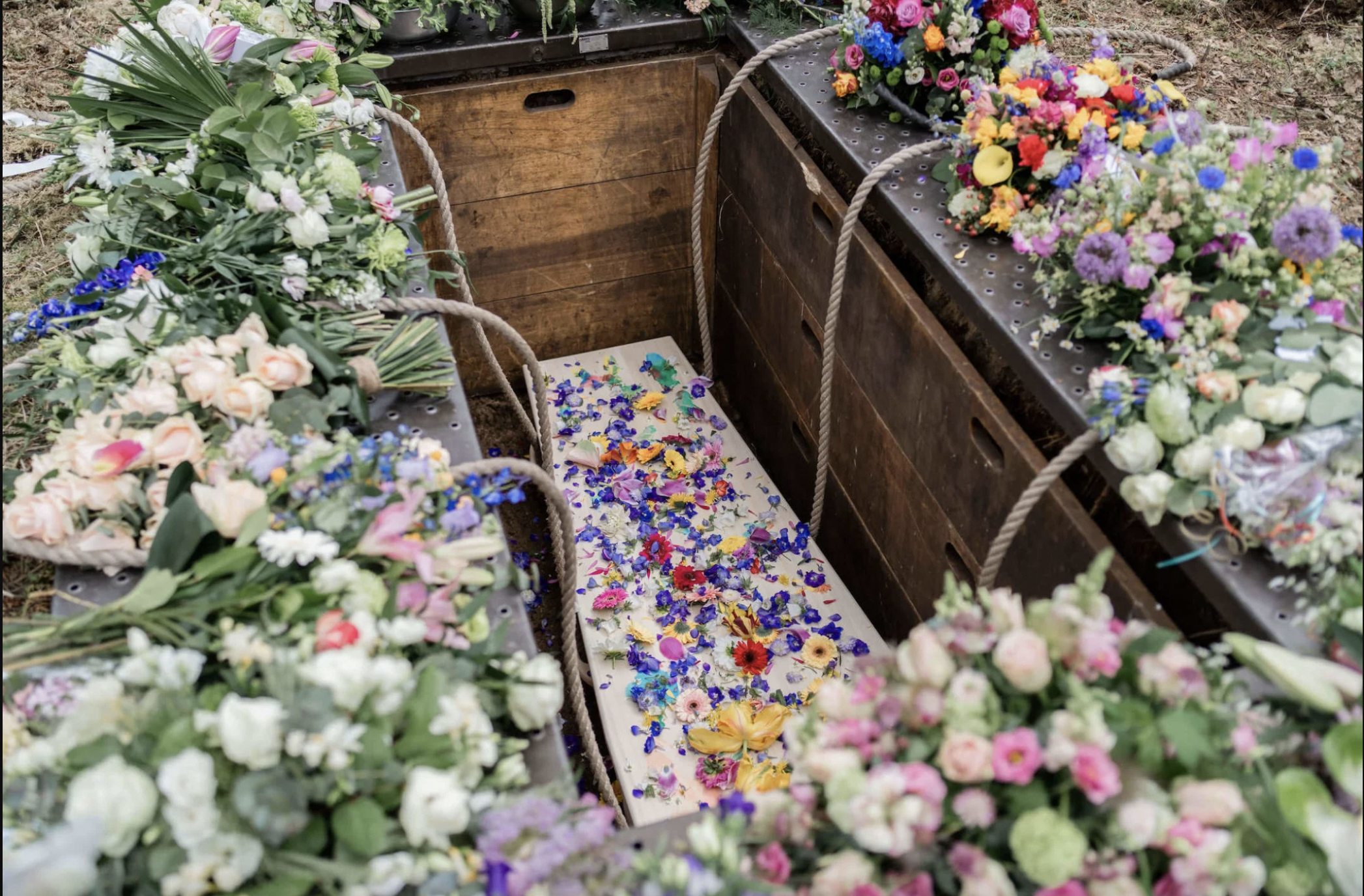 Verschillende bloemen en bloemstukjes, waarschijnlijk voor een begrafenis of gelegenheid, liggen op en rond houten kisten met touwhandgrepen.