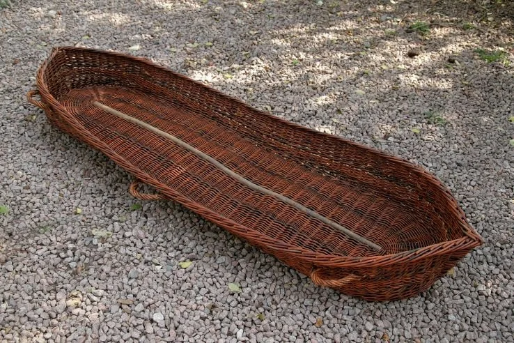 Een langwerpige, lage mand van gevlochten hout, liggend op grind.