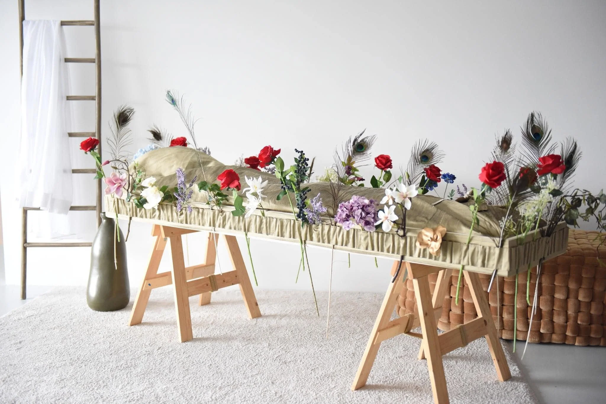 Een opblaasbed met bloemen en veren, geplaatst tussen twee houten tafels in een kamer met een lichte vloer en een witte muur.