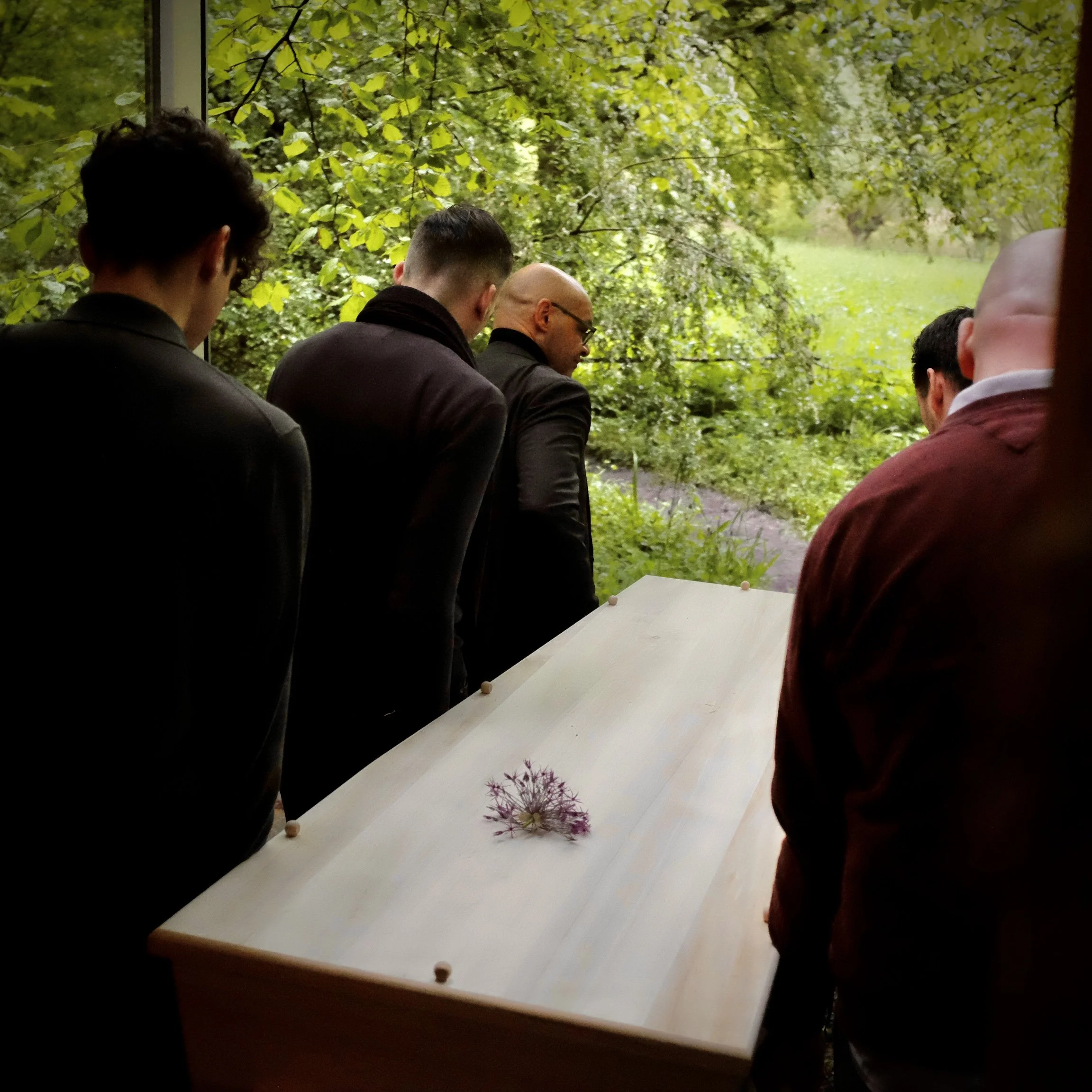 Mensen staan naast een witte kist met een paar bloemen op het deksel, uitkijkend op een groen bos.