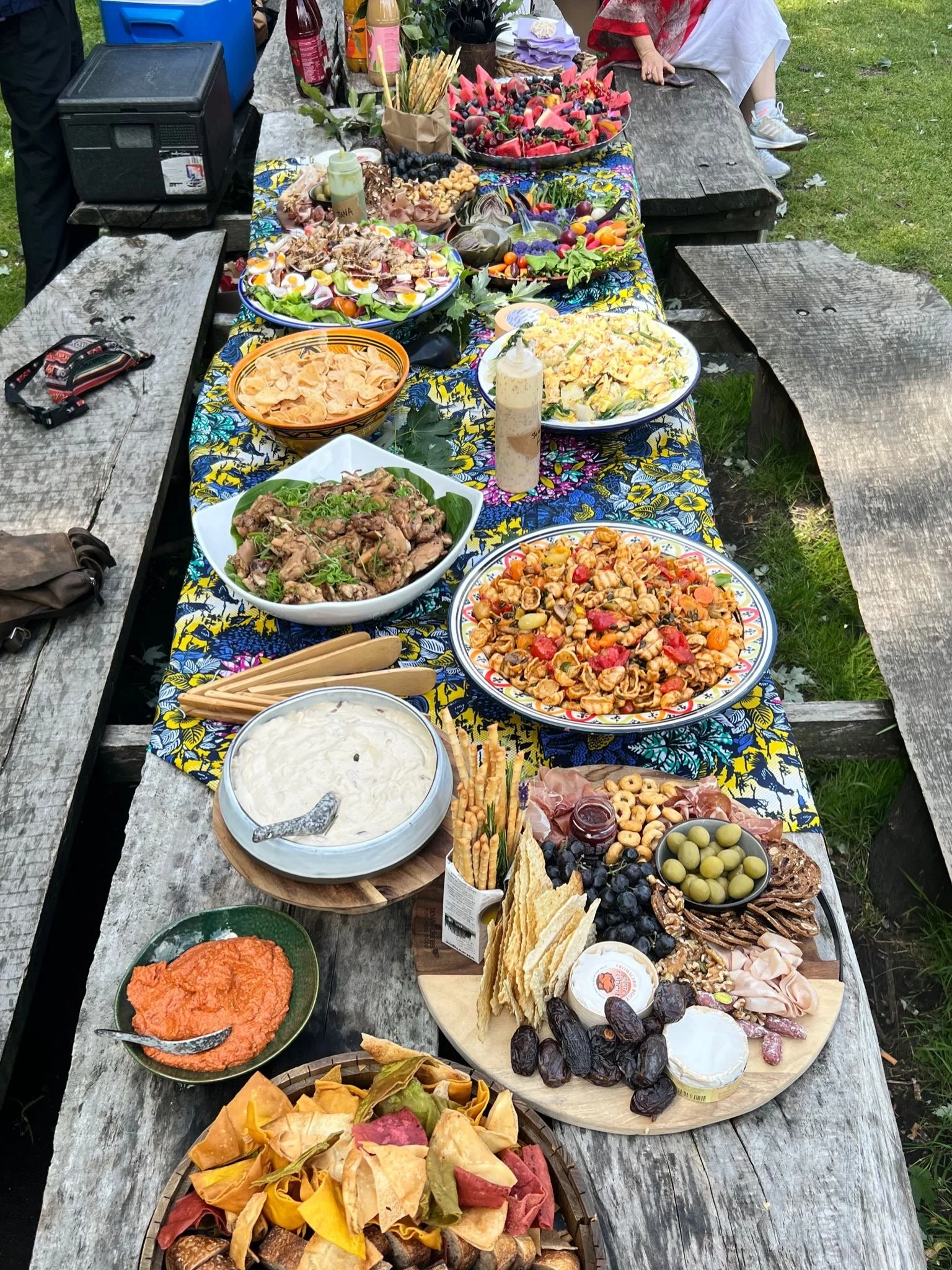 Een lange tafel met diverse gerechten en hapjes, waaronder salade, vlees, olijven, brood en dips, op een buitenlocatie met gras en houten banken.