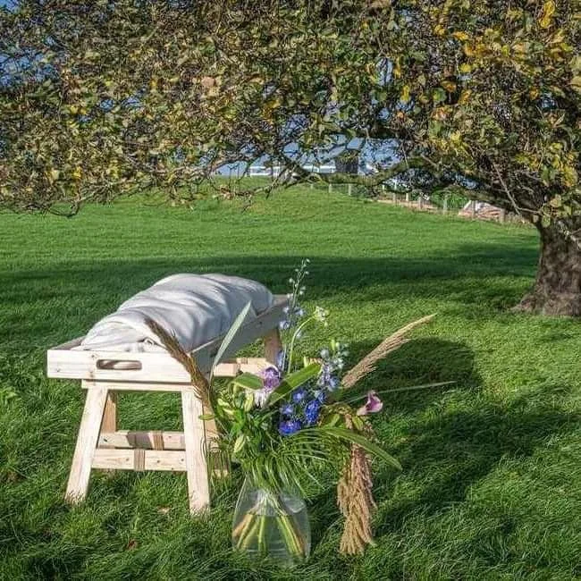 Een bloemenboeket in een vaas, geplaatst voor een houten bank met een witte cloth, onder een boom op een grasveld.