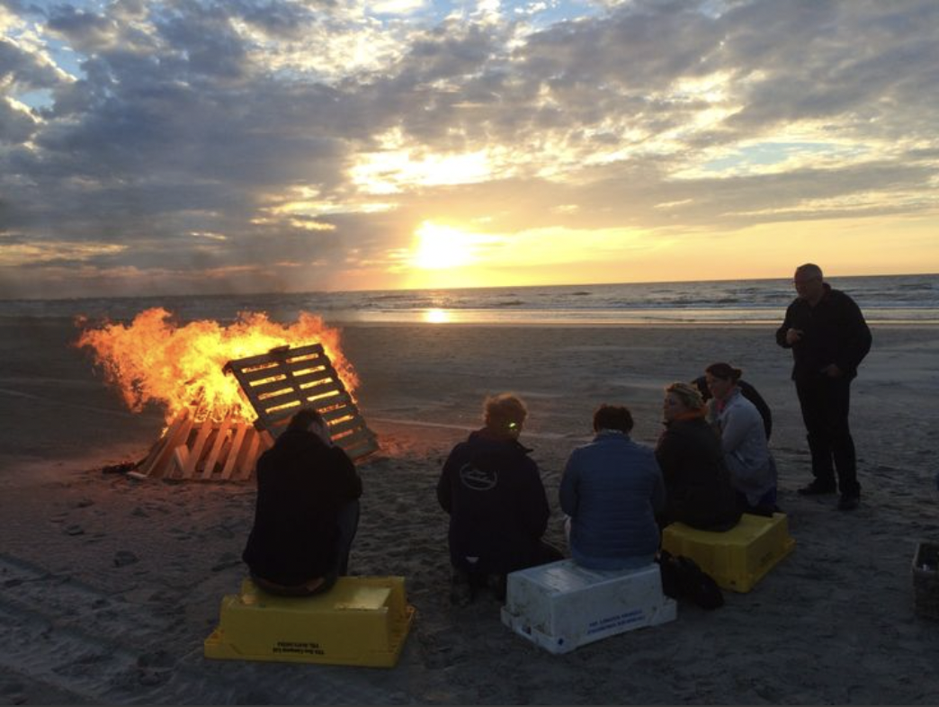 Mensen zitten op plastic bakken op het strand terwijl een kampvuur uit een pallet brandt bij zonsondergang.