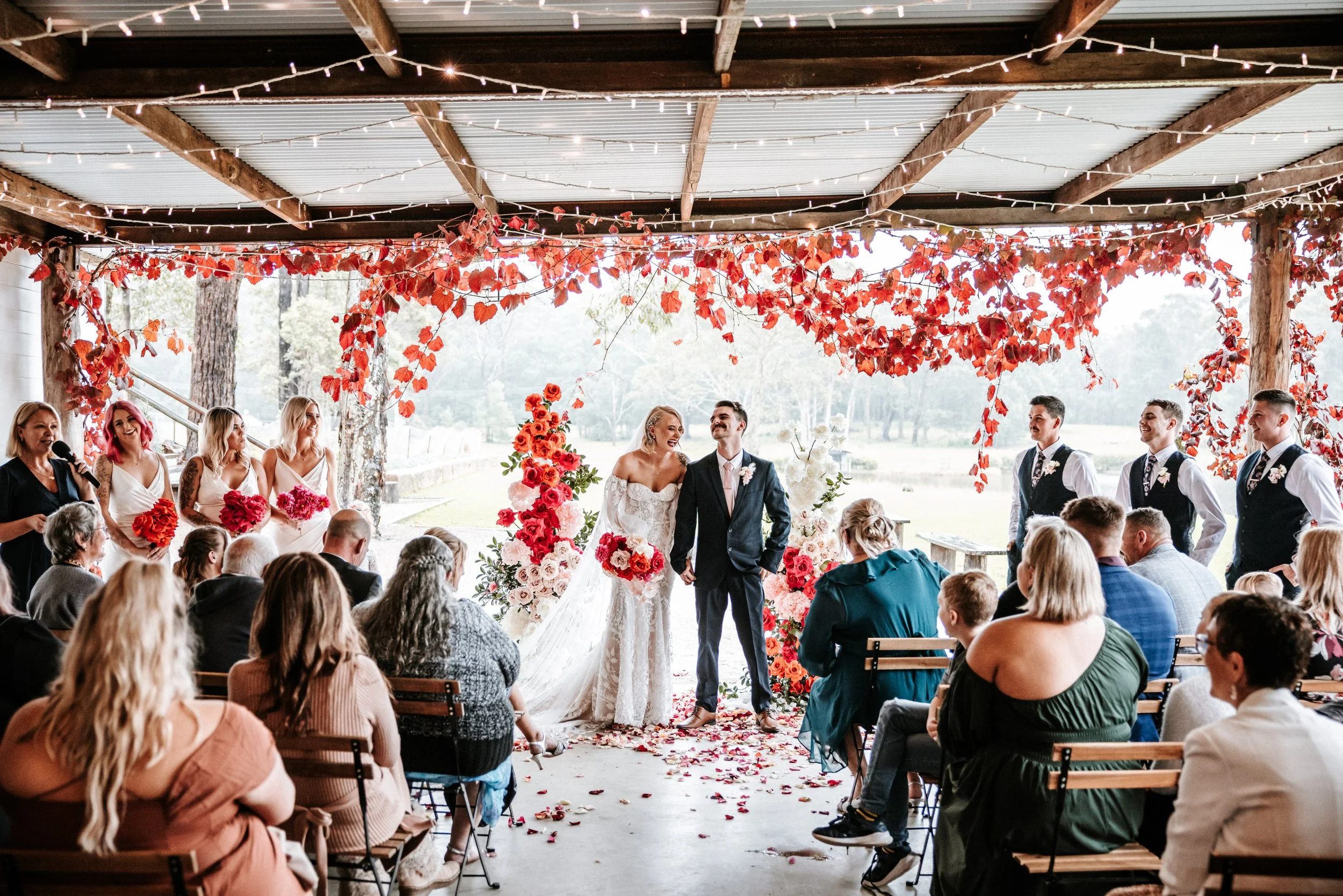 A wedding ceremony taking place outdoors under a decorated shelter with red and pink floral arrangements and leaf garlands. The bride and groom are standing at the altar, smiling, with the bride in a white wedding dress and the groom in a dark suit. Bridesmaids and groomsmen are standing on either side, and guests are seated, watching the ceremony.
