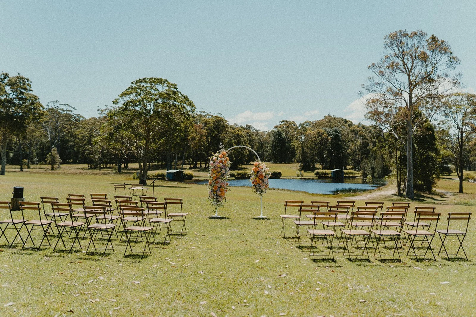 Outdoor wedding ceremony setup on a grassy field with chairs arranged facing a floral arch, near a pond with trees in the background under a blue sky.