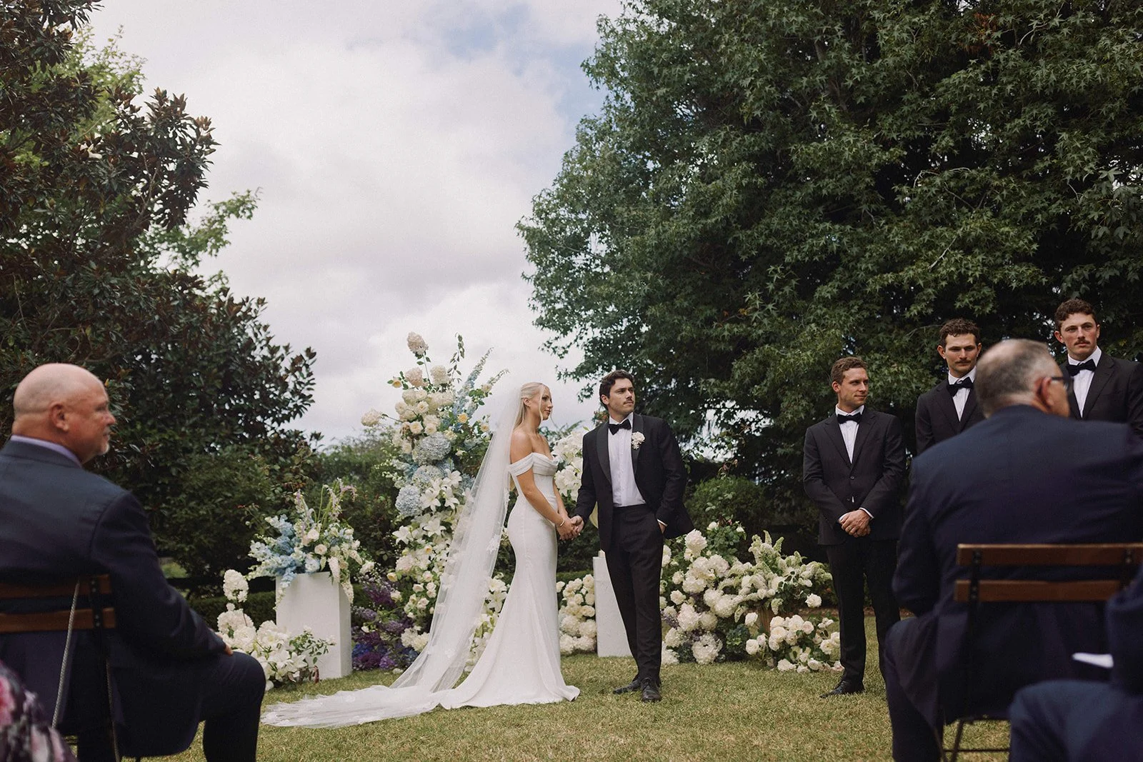 A wedding ceremony outdoors with the bride and groom holding hands, surrounded by floral arrangements, with groomsmen and guests nearby under a cloudy sky.
