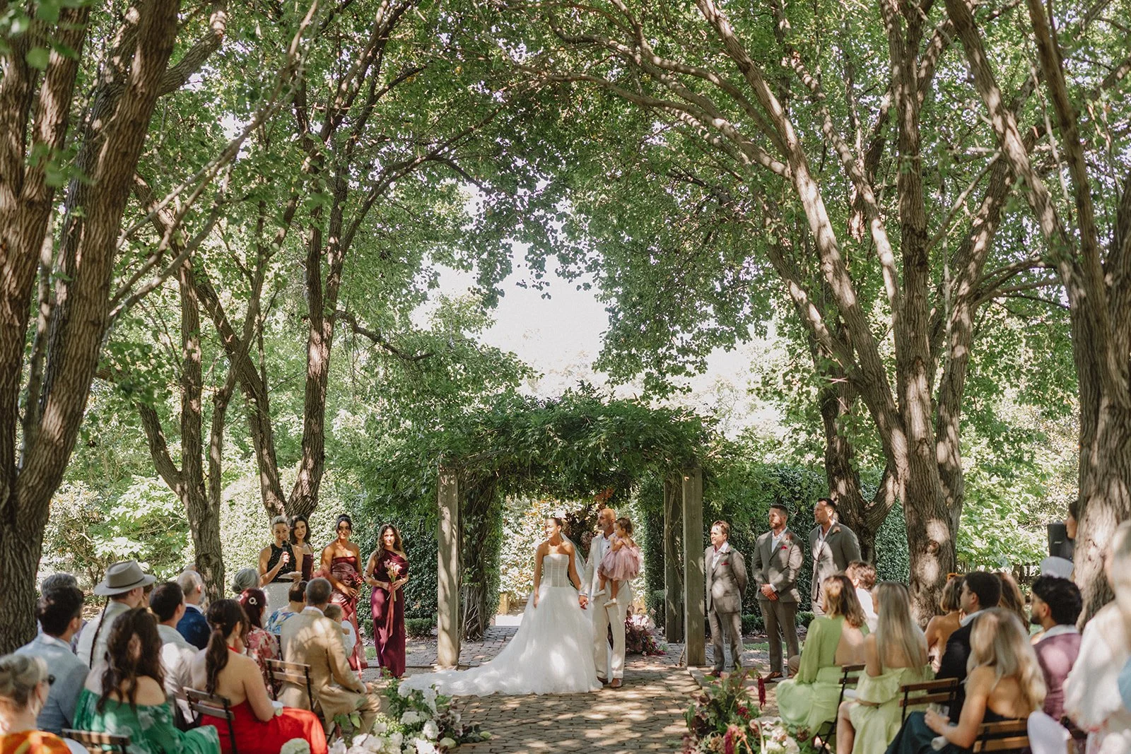 Wedding ceremony outdoors under large trees with a bride and groom holding hands, surrounded by guests and wedding party, wedding arch decorated with greenery.