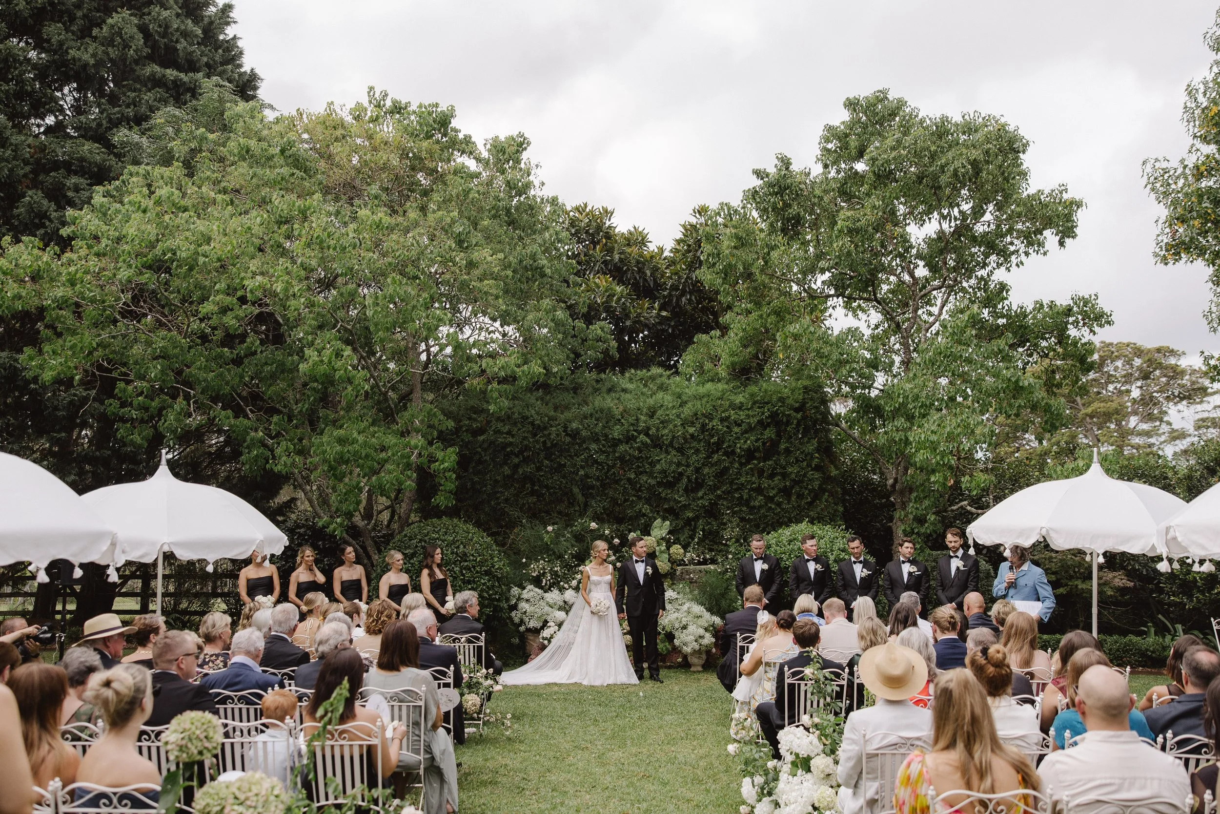 Outdoor wedding setup in a garden with white chairs, white umbrellas, and floral arrangements of white and pink flowers lining the aisle, surrounded by green trees and bushes.