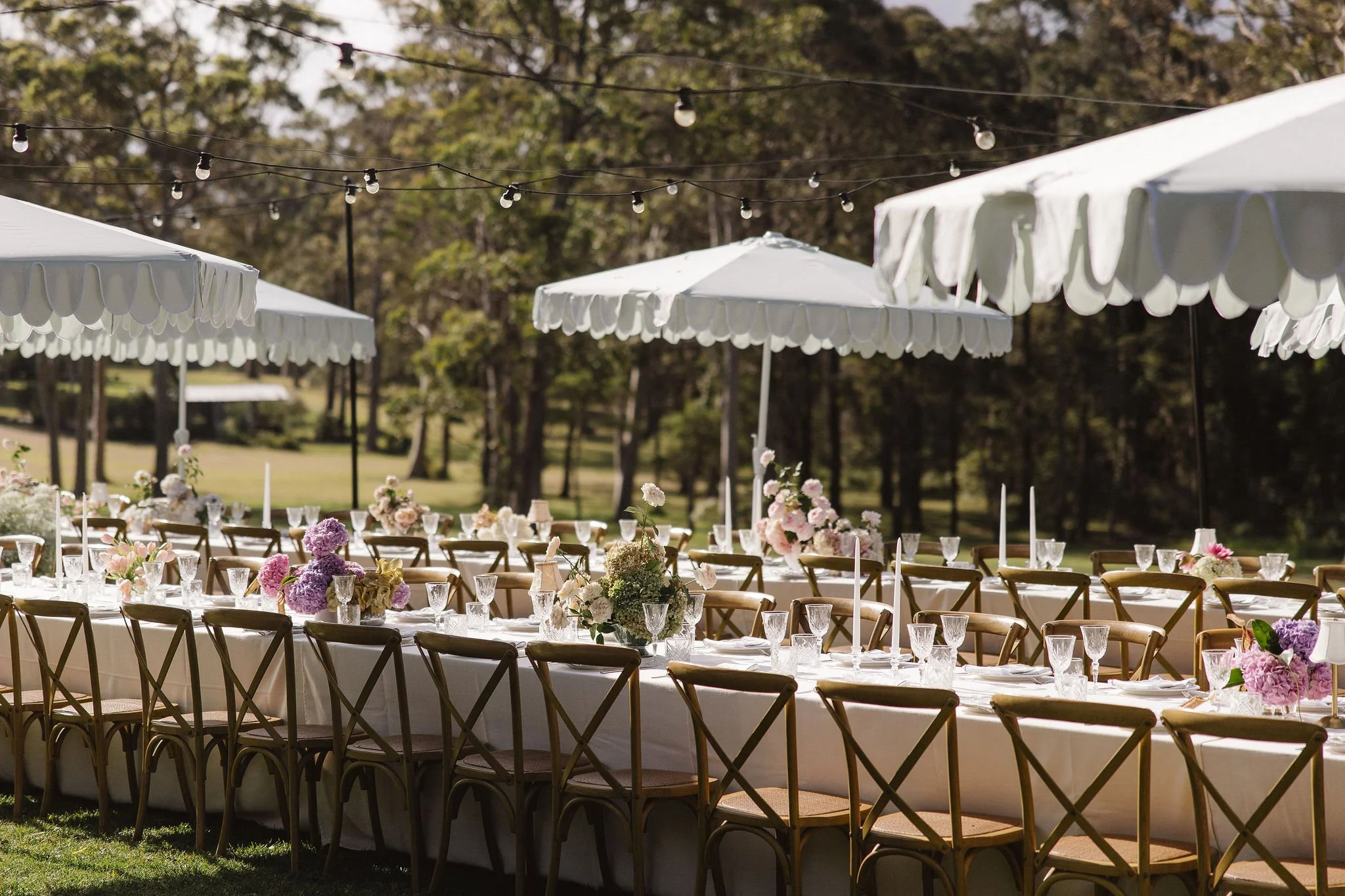 Outdoor wedding reception setup with long tables, white tablecloths, floral centerpieces in pink, purple, and white, glassware, and white umbrellas providing shade, surrounded by wooden chairs in a grassy area with trees in the background.