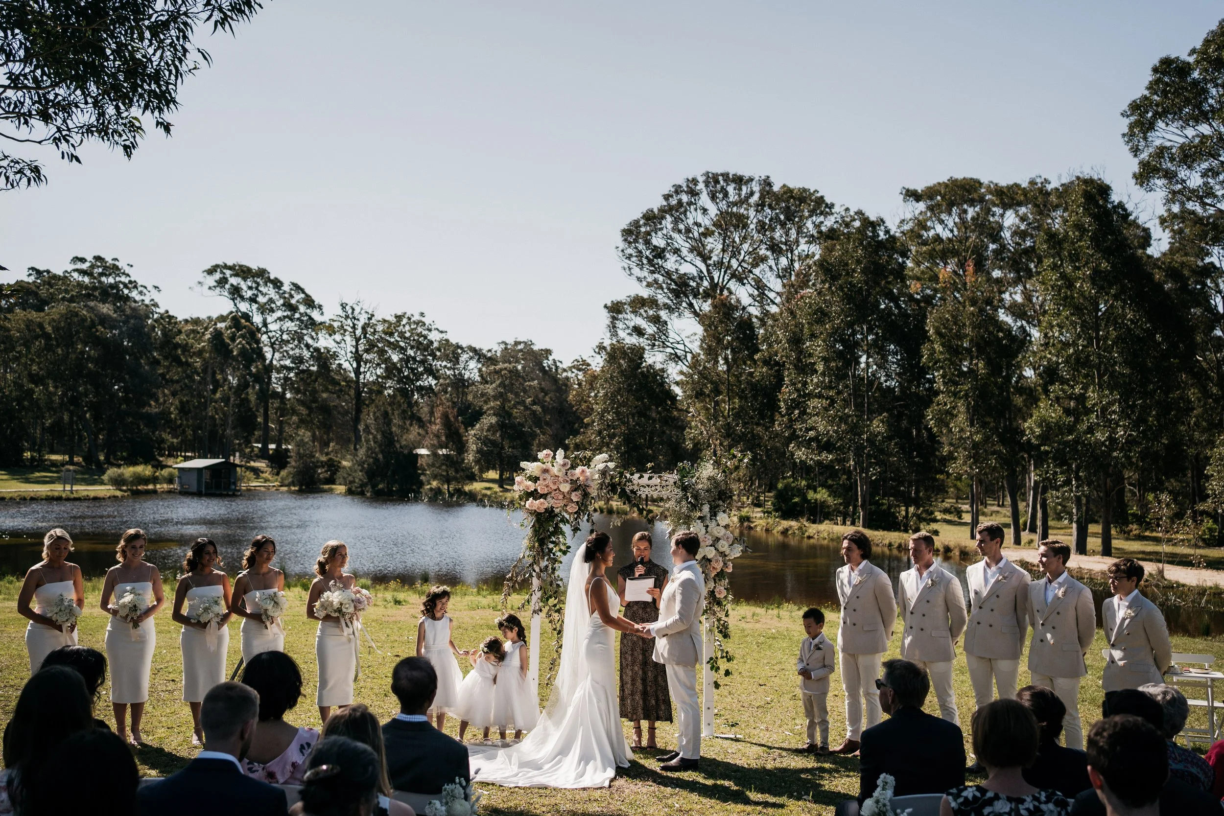 A wedding ceremony outdoors by a lake with trees in the background. The bride and groom are holding hands and exchanging vows under a floral arch, with bridesmaids and groomsmen standing on either side. Guests are seated and watching the ceremony.