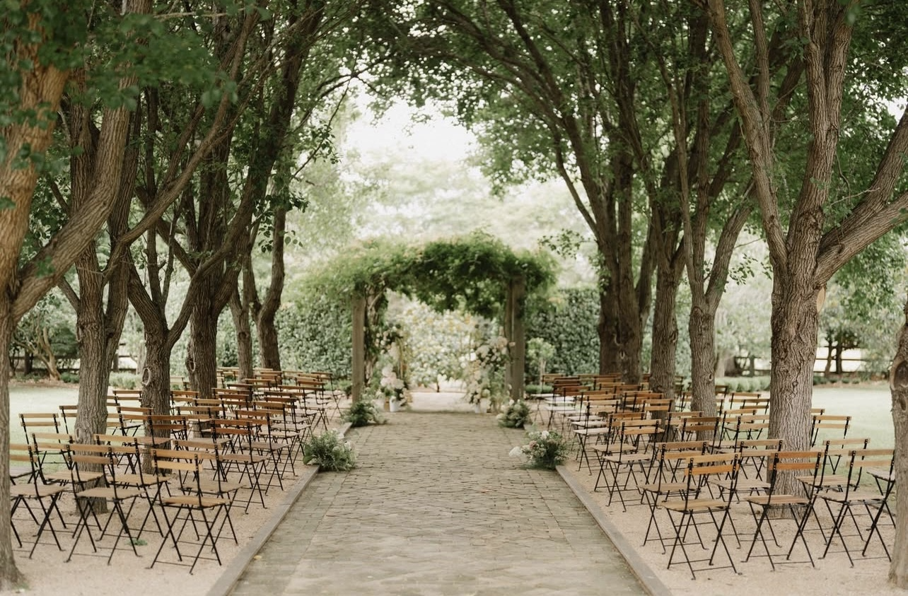 Outdoor wedding ceremony setup with a brick path, rows of wooden chairs, surrounded by trees, and an altar decorated with flowers and greenery.