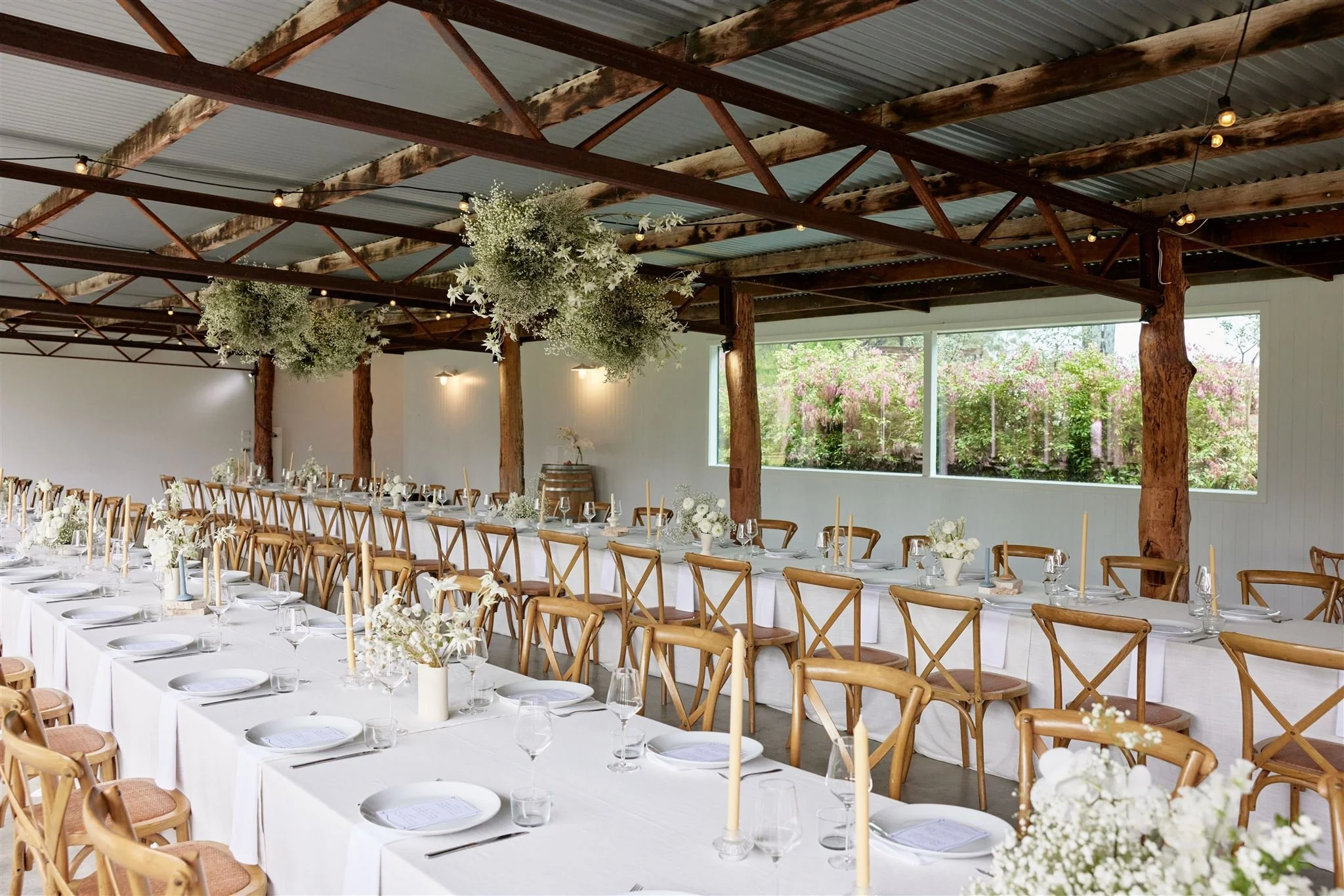 Long banquet tables set with white tablecloths, white plates, glassware, and floral centerpieces in an airy rustic event space with wooden beams, hanging greenery, and large windows showing pink flowering bushes outside.