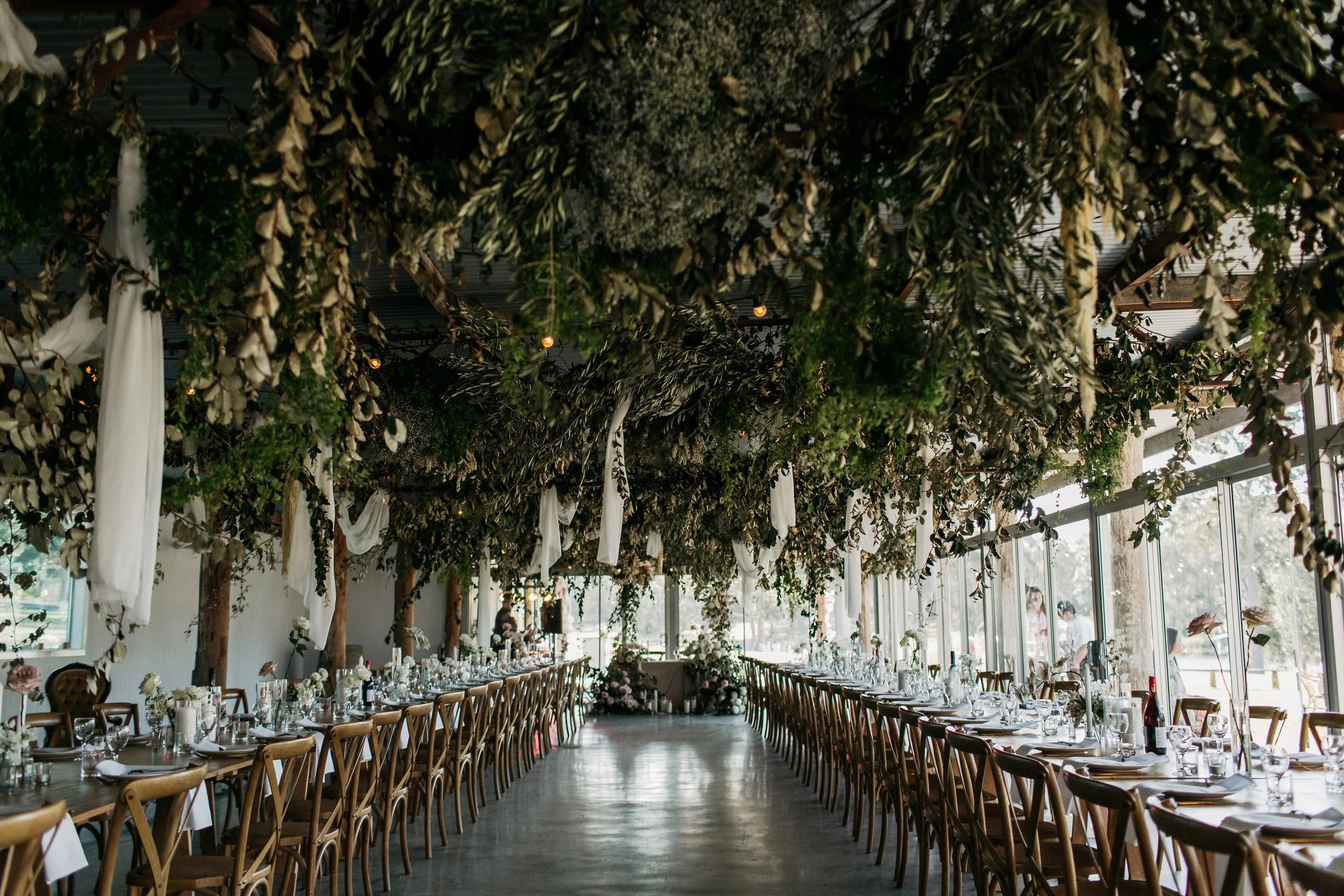 A banquet hall decorated with hanging greenery and white drapes, with long tables set for a formal event, featuring floral centerpieces, glassware, and place settings, illuminated by natural light from large windows.