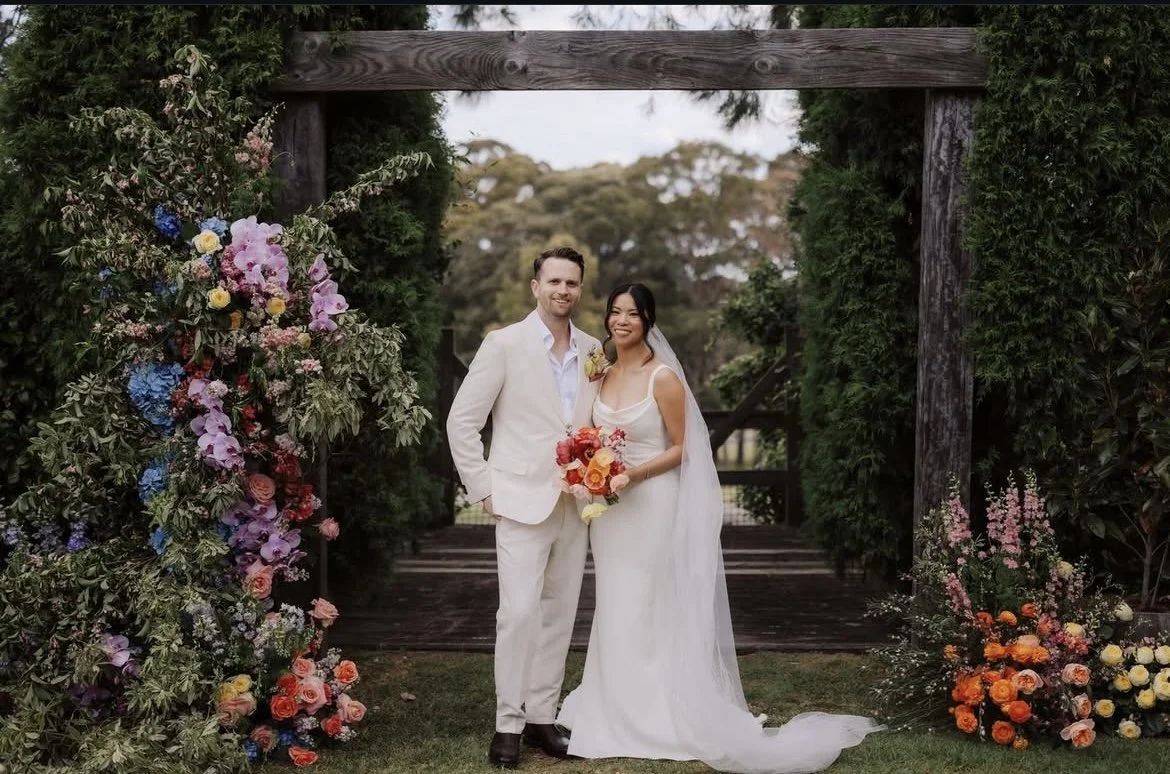 A newlywed couple stands together outdoors in front of a floral arch and greenery, holding a bouquet of pink and orange flowers. The groom has light skin, dark hair, a white suit, and is smiling, while the bride has light skin, dark hair, a white wedding dress, and is also smiling.