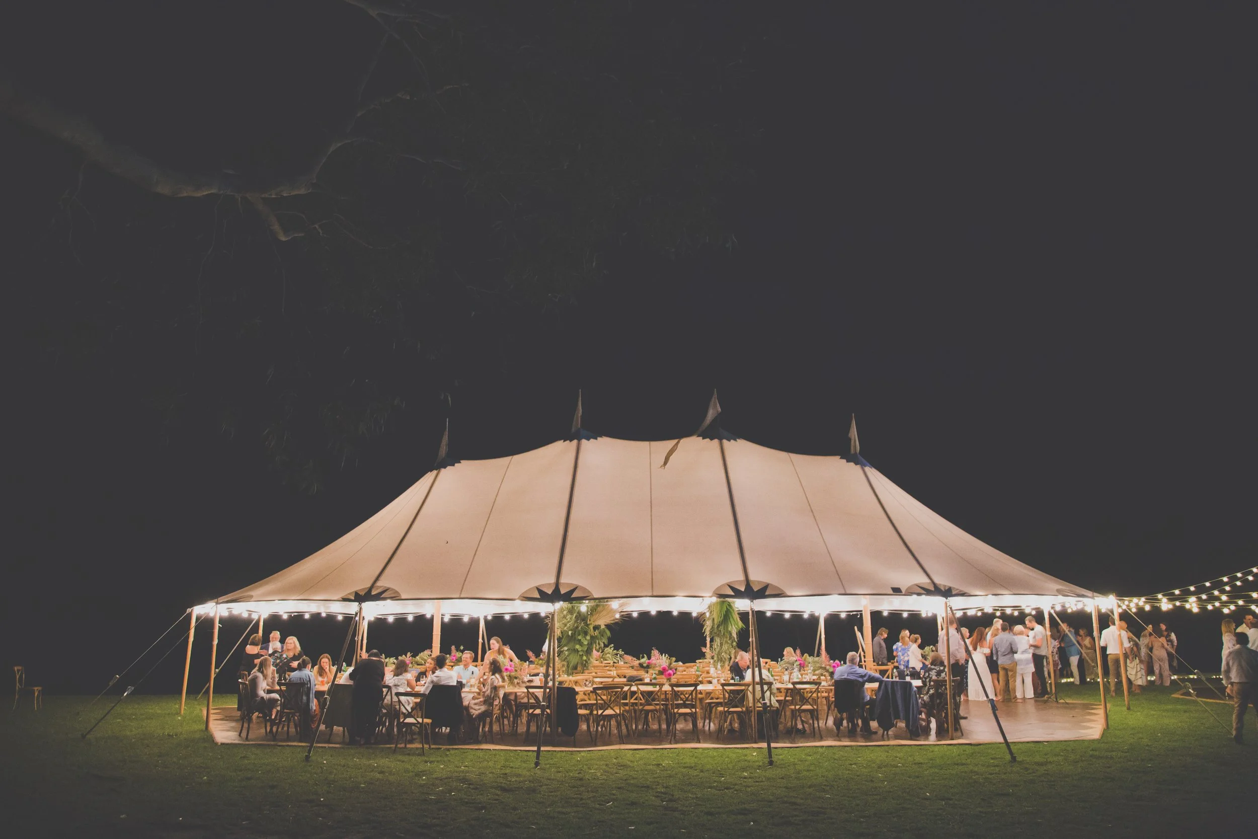 Nighttime outdoor wedding reception under a large tent with string lights, decorated tables, and people socializing.