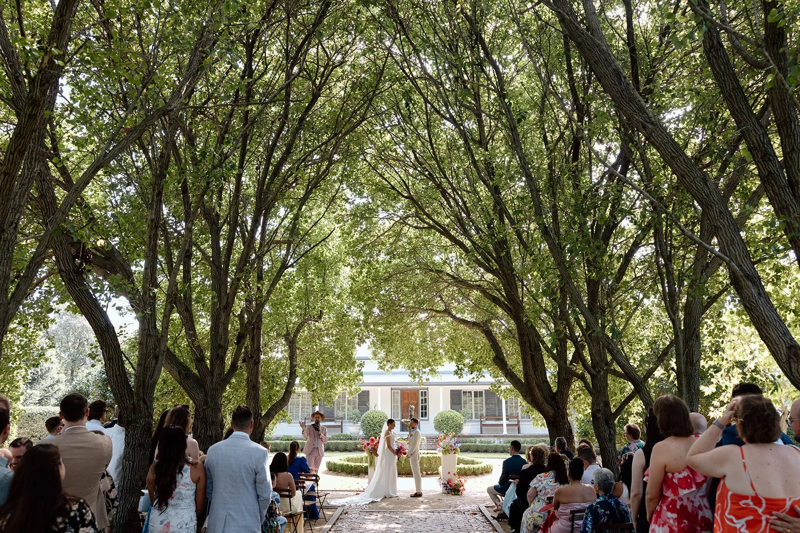 Wedding ceremony outdoors under large leafy trees, with a bride and groom facing each other, surrounded by seated guests and an officiant.