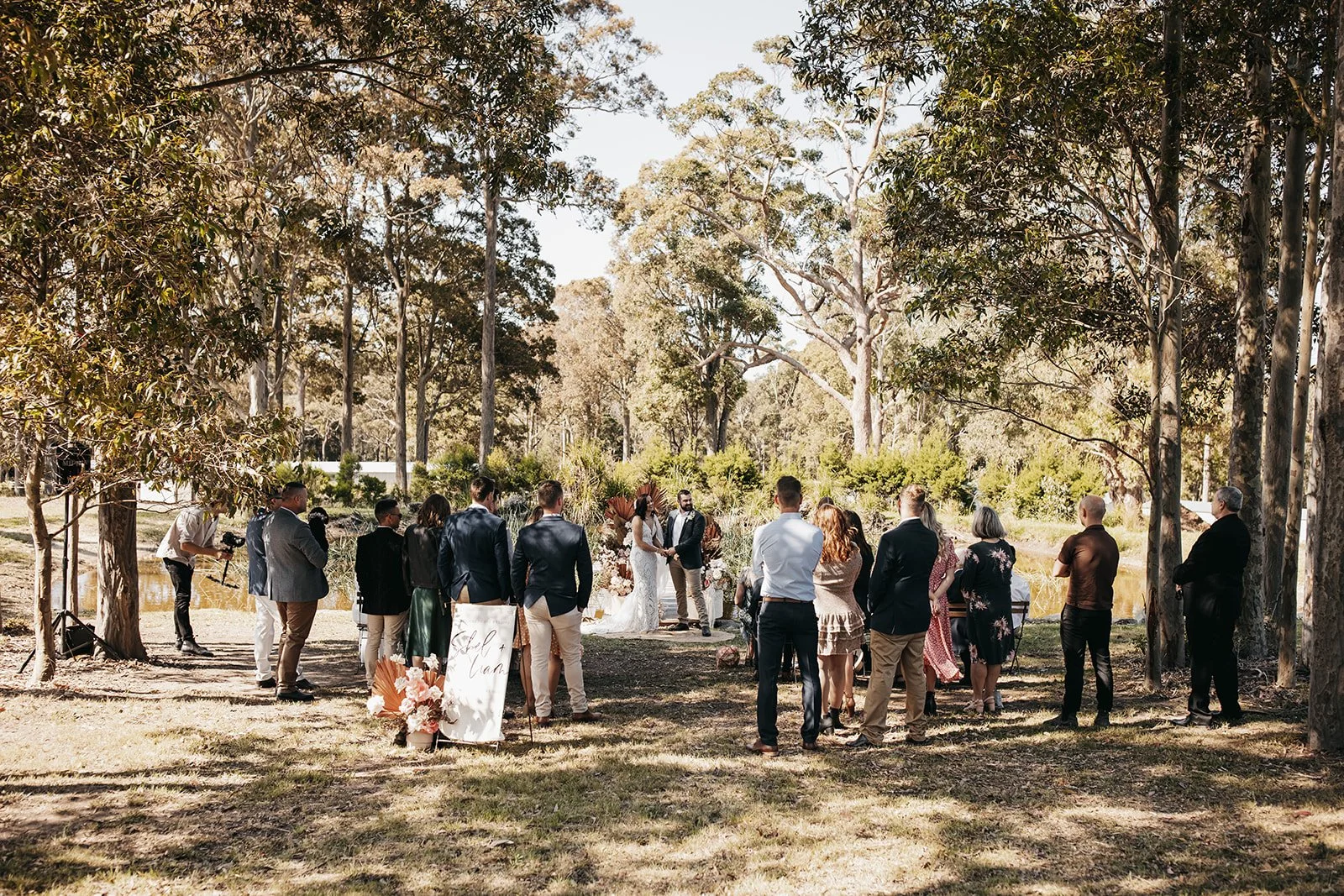 A wedding ceremony taking place outdoors in a wooded park with trees and a pond in the background. The bride and groom stand in front of the wedding party, surrounded by guests, under a partly shaded area on a sunny day.